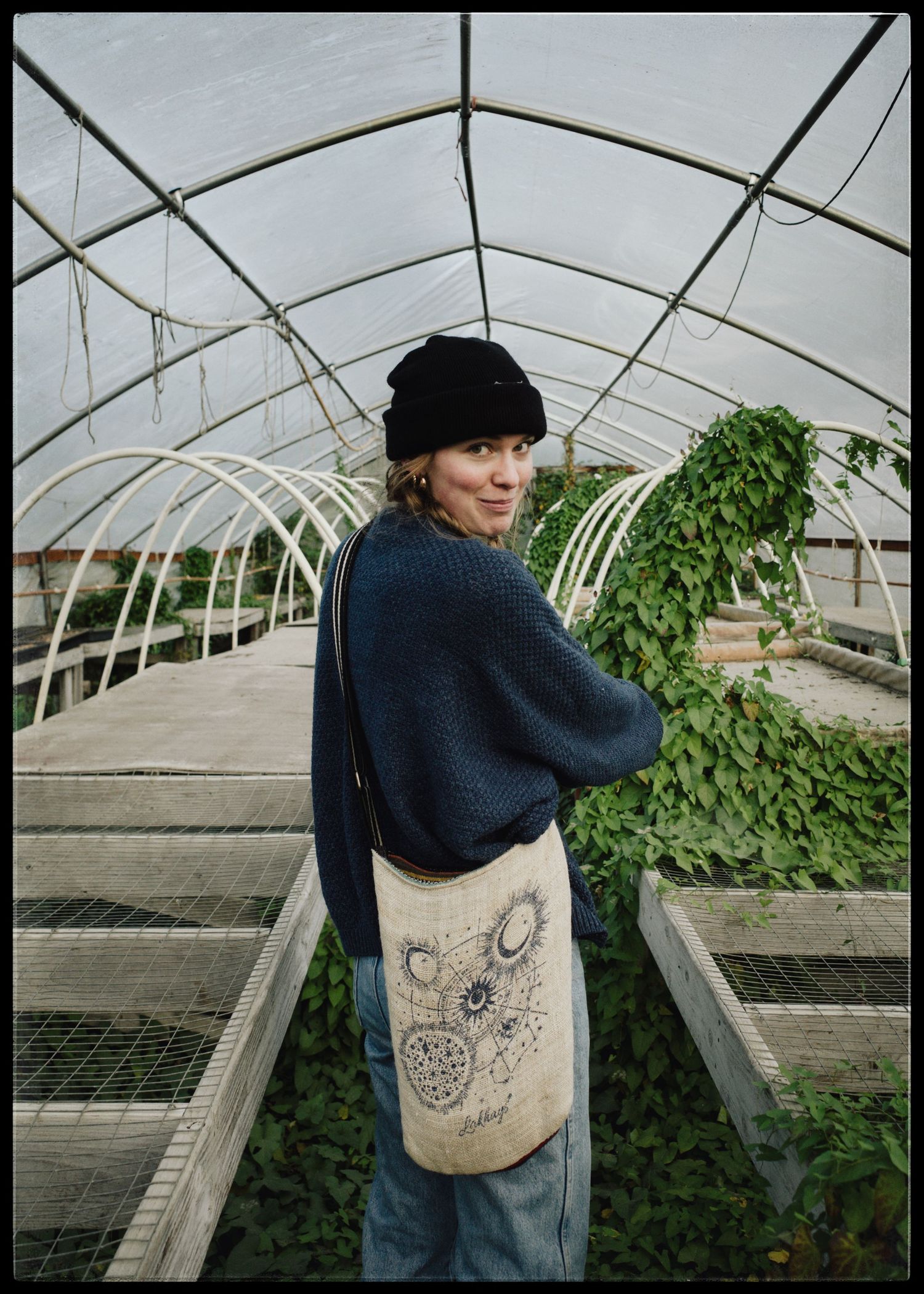 Woman in black beanie and blue sweater holding a tote bag stands in a greenhouse with lush green plants.