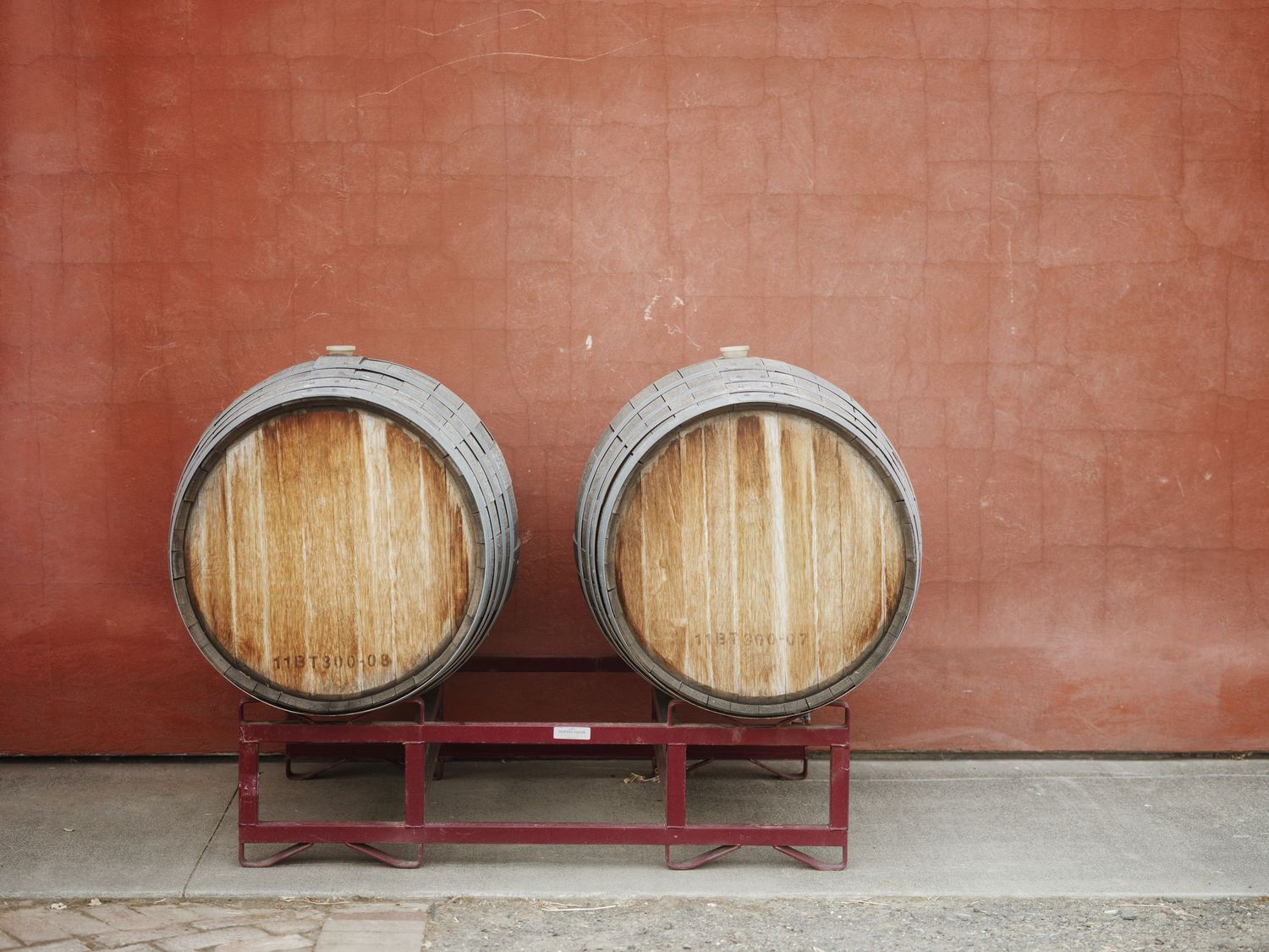 Two wooden wine barrels on a red metal rack against a terracotta brick wall outdoors.