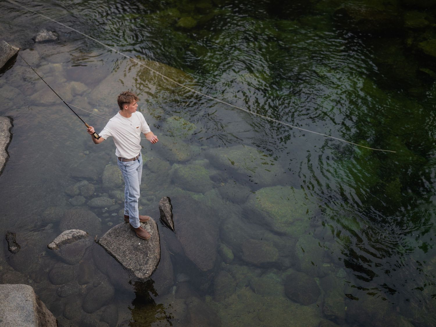 Man standing on rocks fly fishing in a clear green river with visible stones underwater.