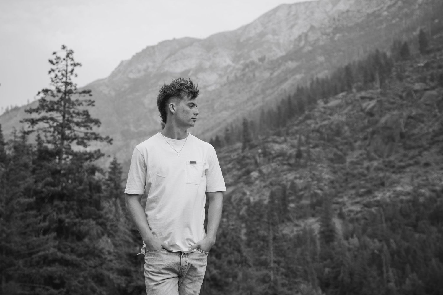Young man in white t-shirt stands before misty mountain forest landscape in black and white photo.