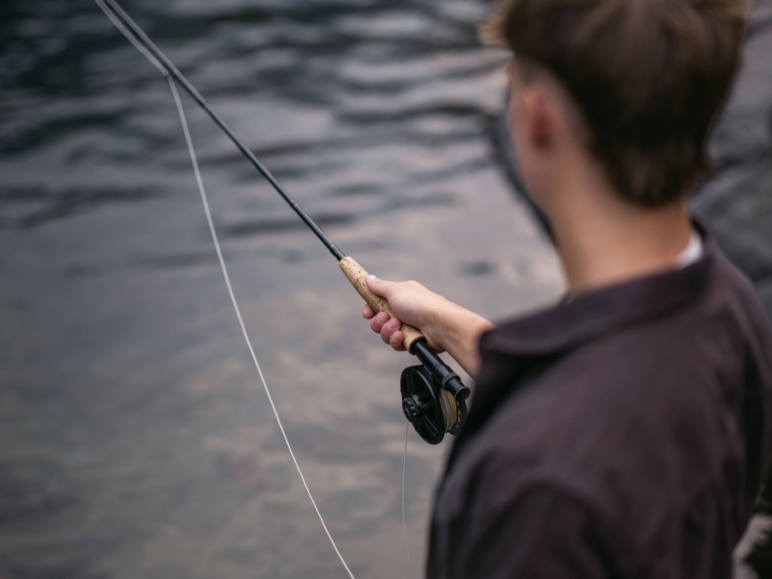 Young man fly fishing by a river, holding a fishing rod with reel over calm dark water.