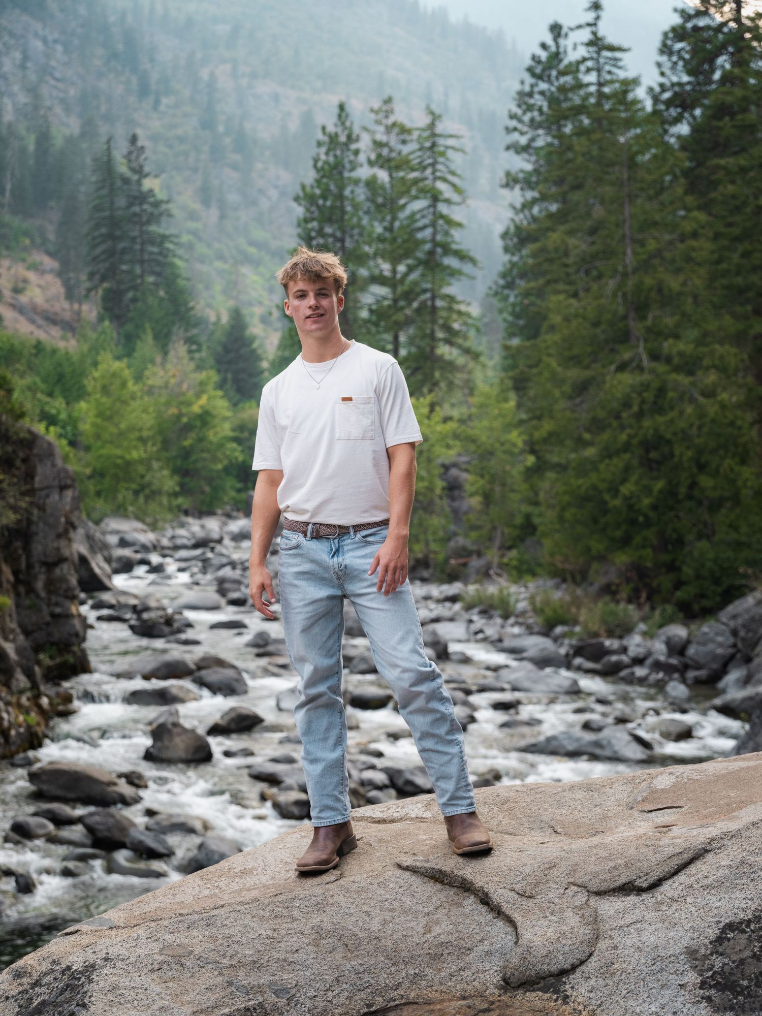 Young man in white tee and jeans standing on rocks by a mountain stream surrounded by evergreen trees.
