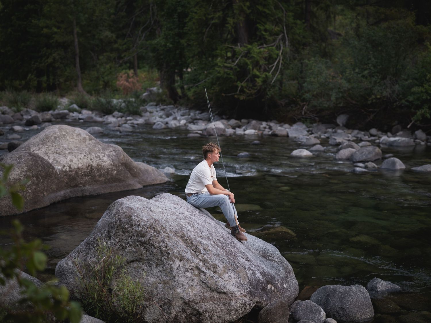 Person sitting alone on large boulder by a rocky river surrounded by lush green forest trees.