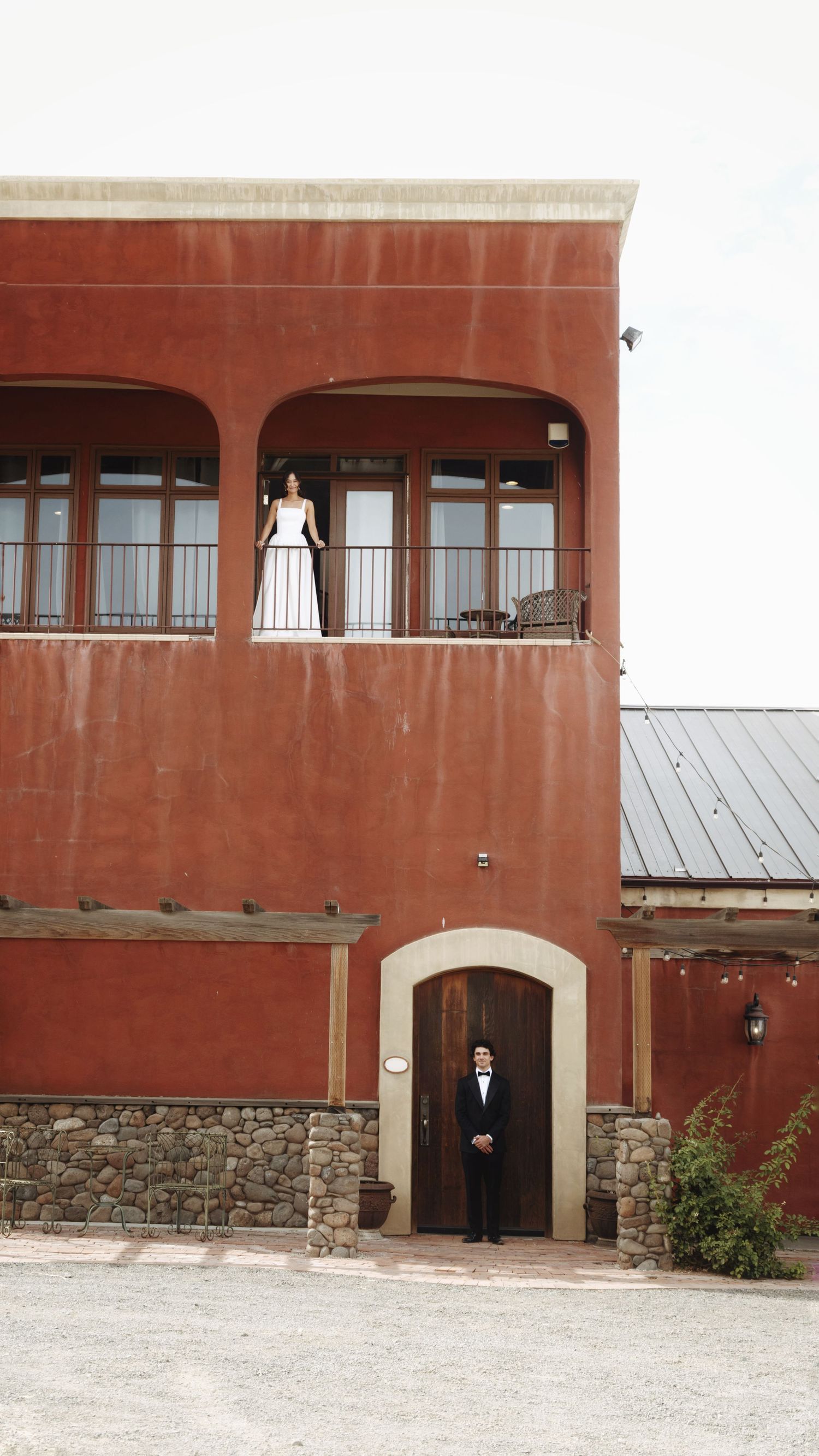 Bride on balcony and groom at entrance of rustic red stucco wedding venue building.