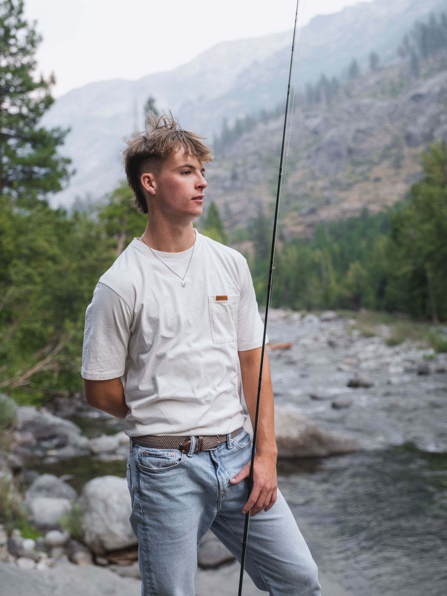 Young man holding a fishing rod by a mountain river, wearing white t-shirt and jeans, with forested hills behind.