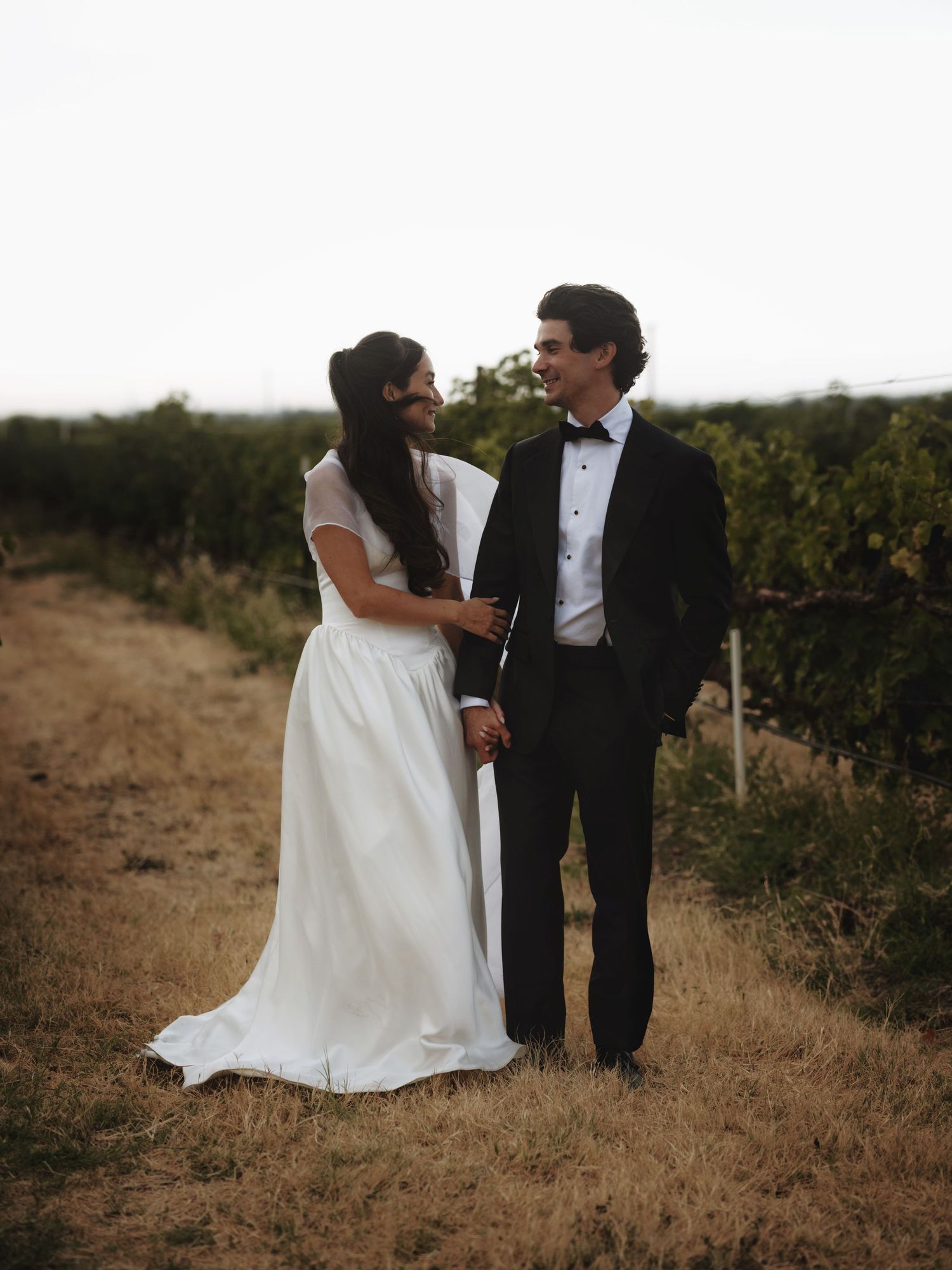 A bride in white gown and groom in black tuxedo hold hands in a vineyard field at dusk.