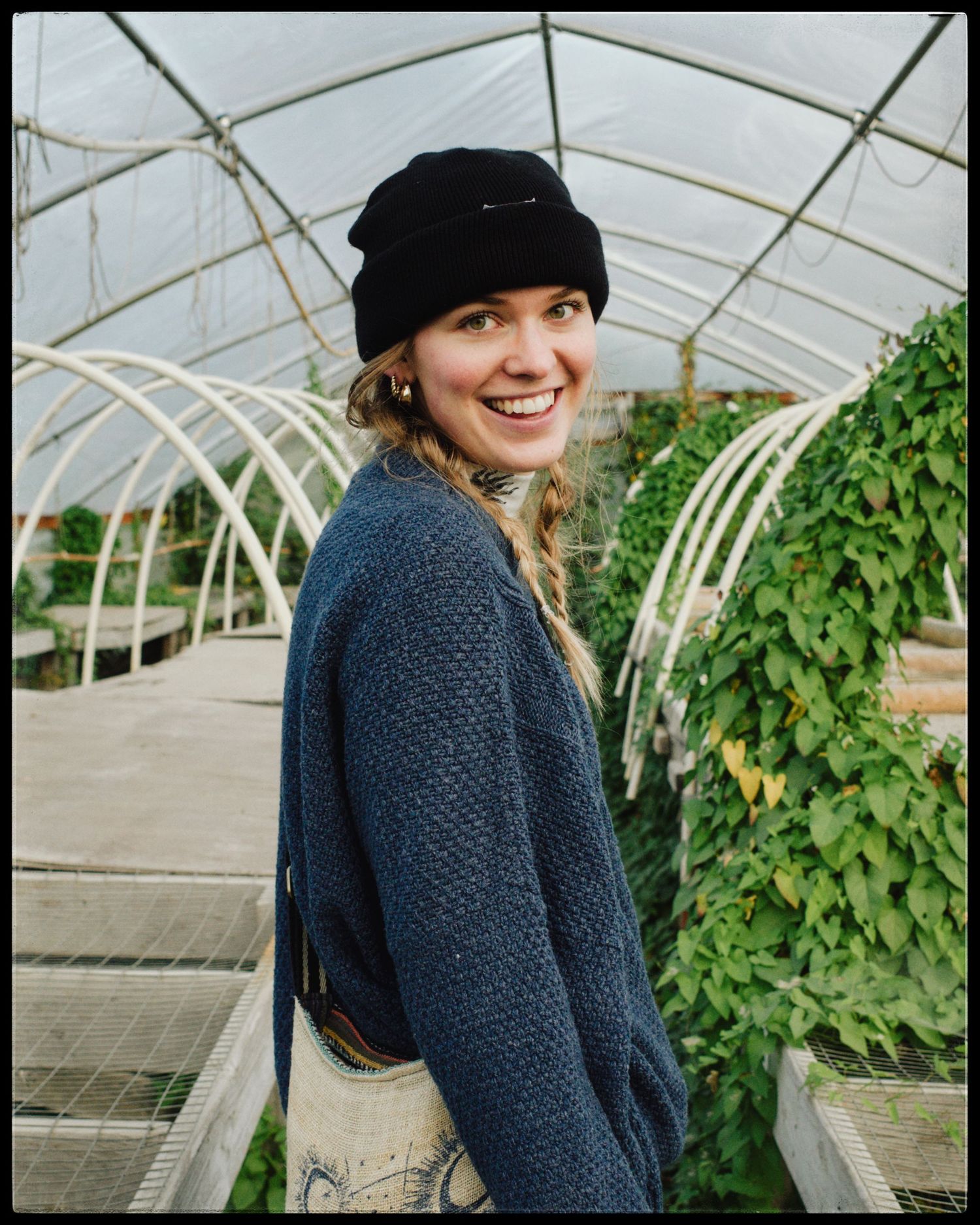 Smiling woman in black beanie and blue knit sweater standing in a lush greenhouse with green plants.