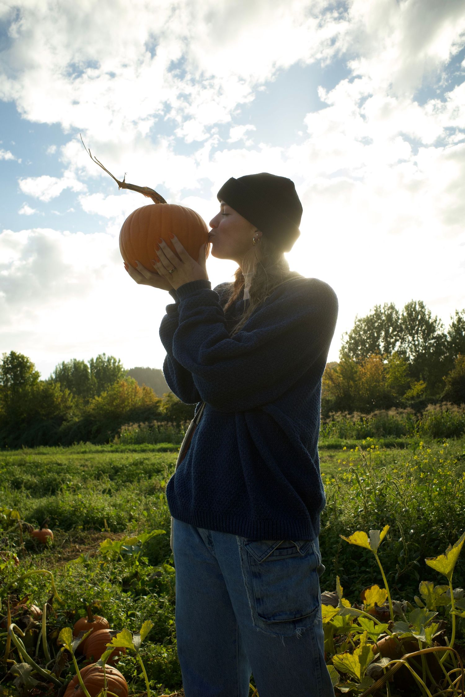 Young woman in beret kissing a pumpkin in a sunny autumn field, wearing blue sweater and jeans.