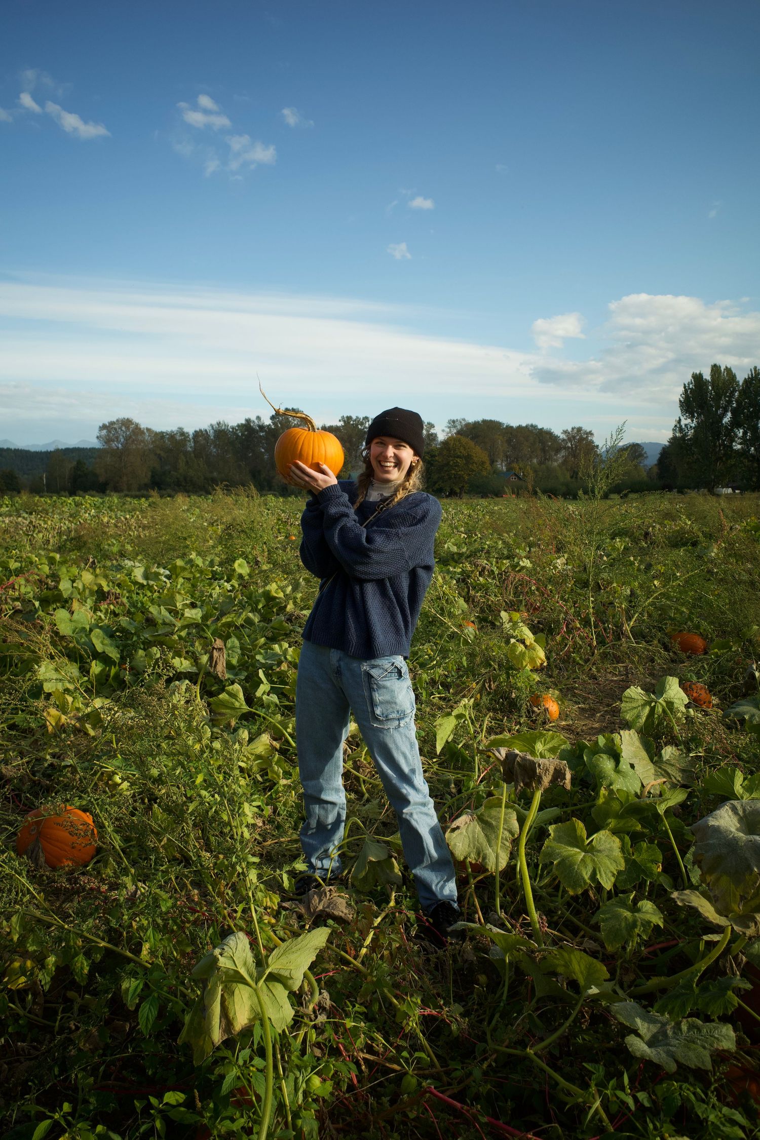Young woman smiling in a pumpkin patch, holding an orange pumpkin up on a sunny autumn day.