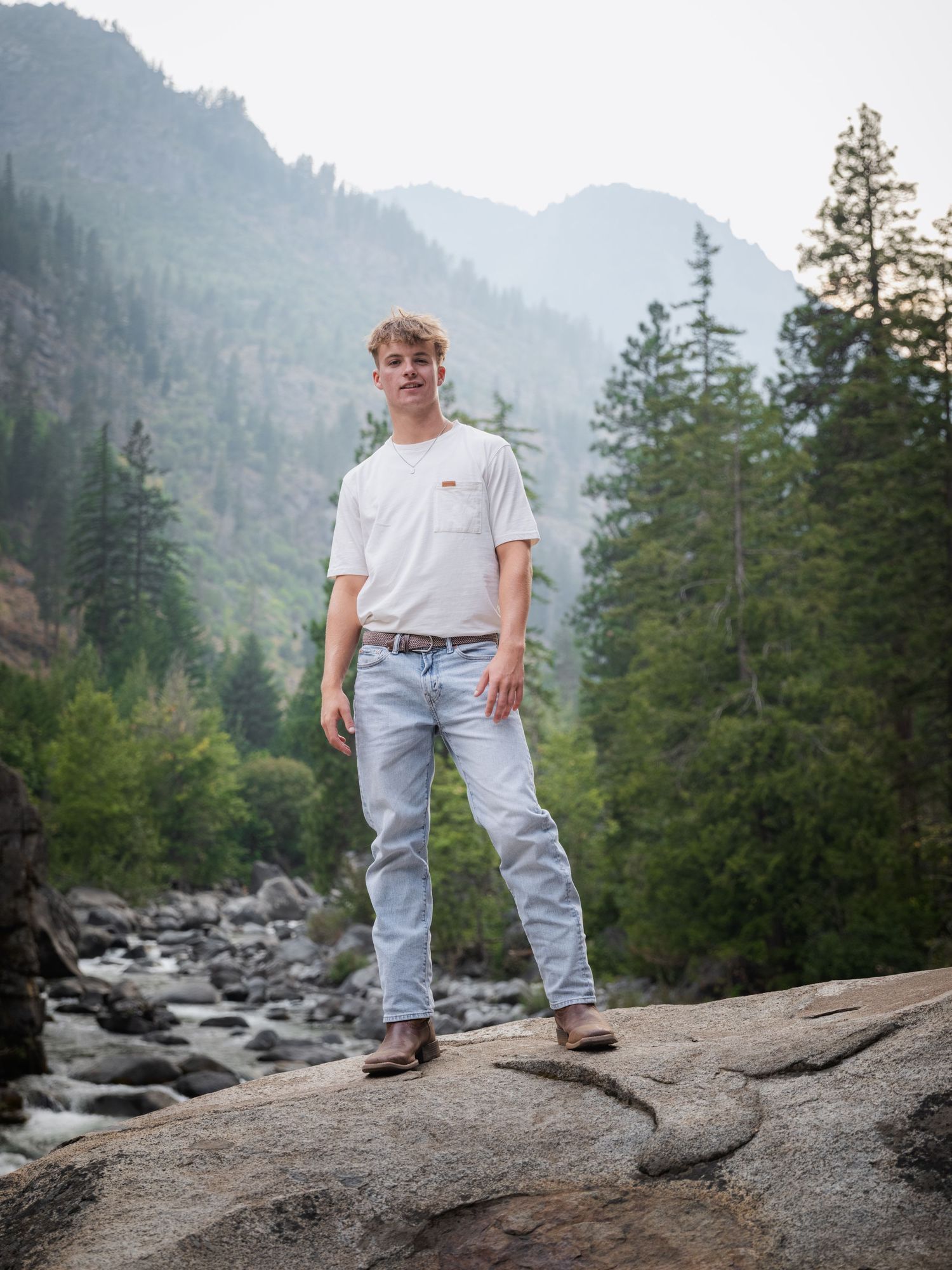 Young man in white tee and light jeans stands on rocks with misty mountain forest backdrop.