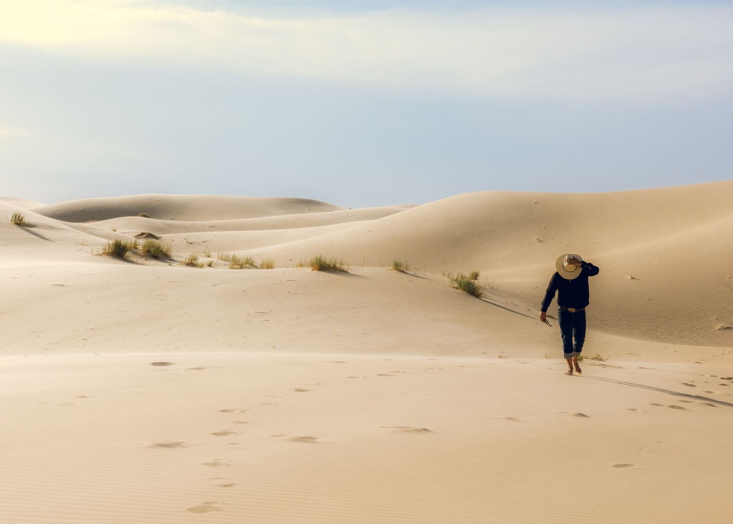 Person hiking alone across vast desert sand dunes under cloudy sky.