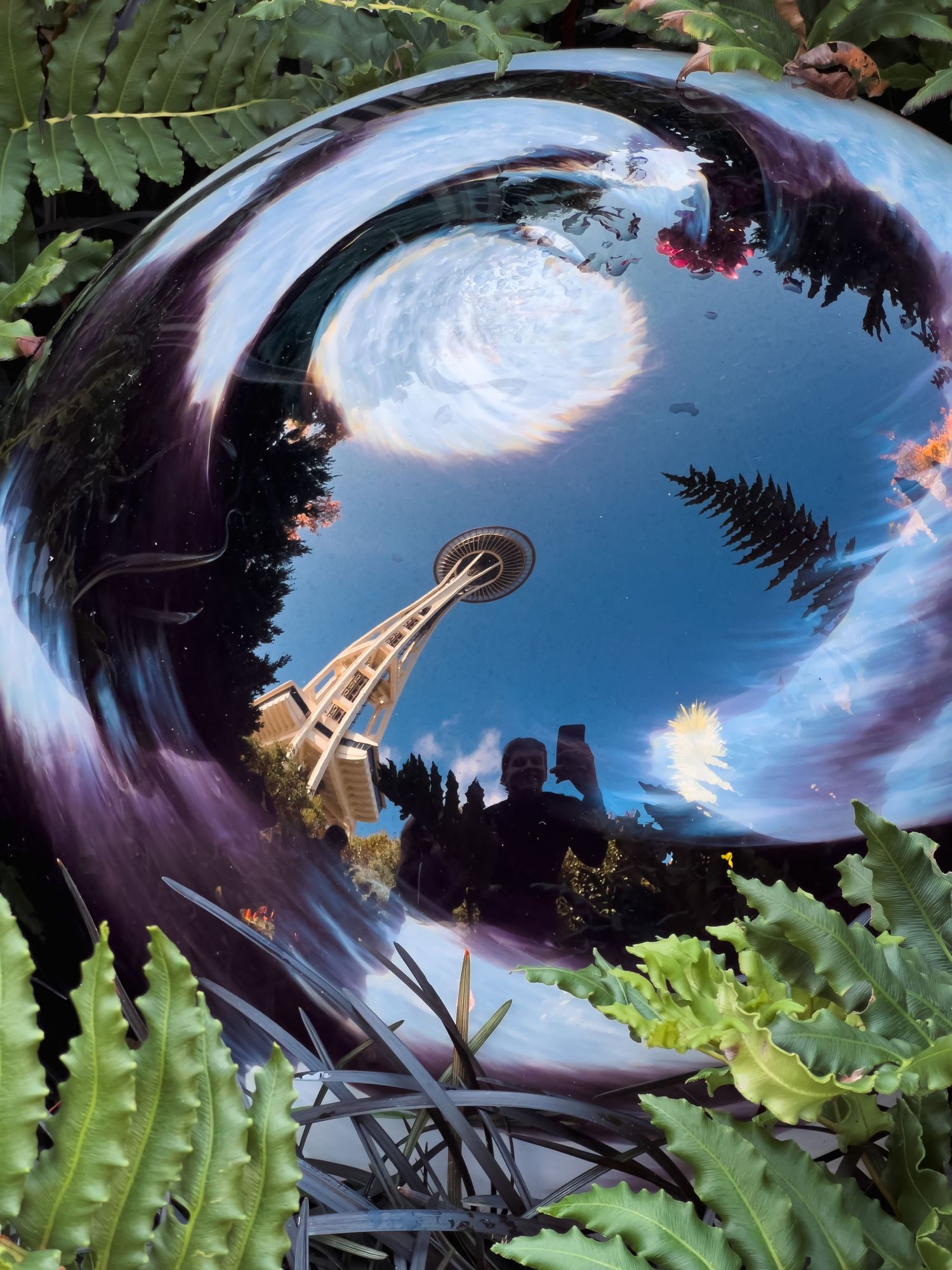 Artistic reflection of the Space Needle in a curved metallic surface surrounded by foliage.
