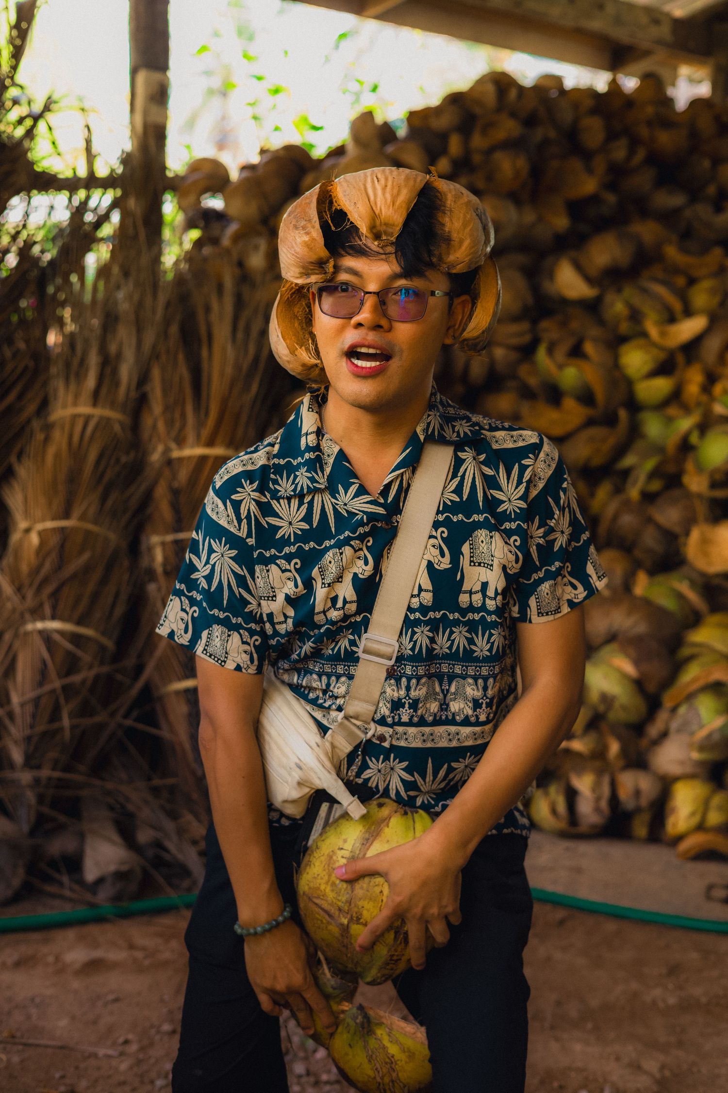 Young man in elephant print shirt and coconut hat holds fresh coconuts at a tropical farm market.
