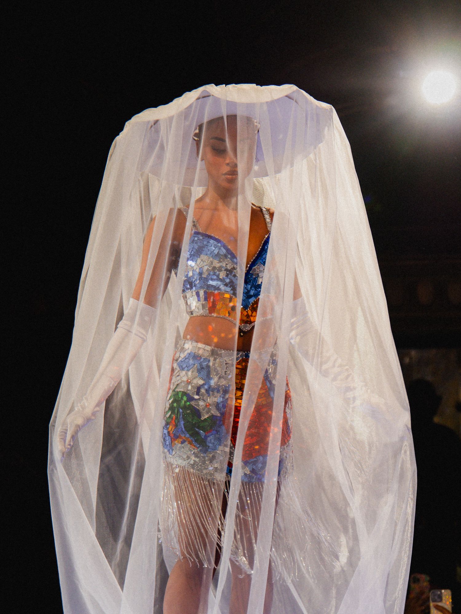 Model walks runway in sheer white overlay dress with floral blue pattern underneath during fashion show.