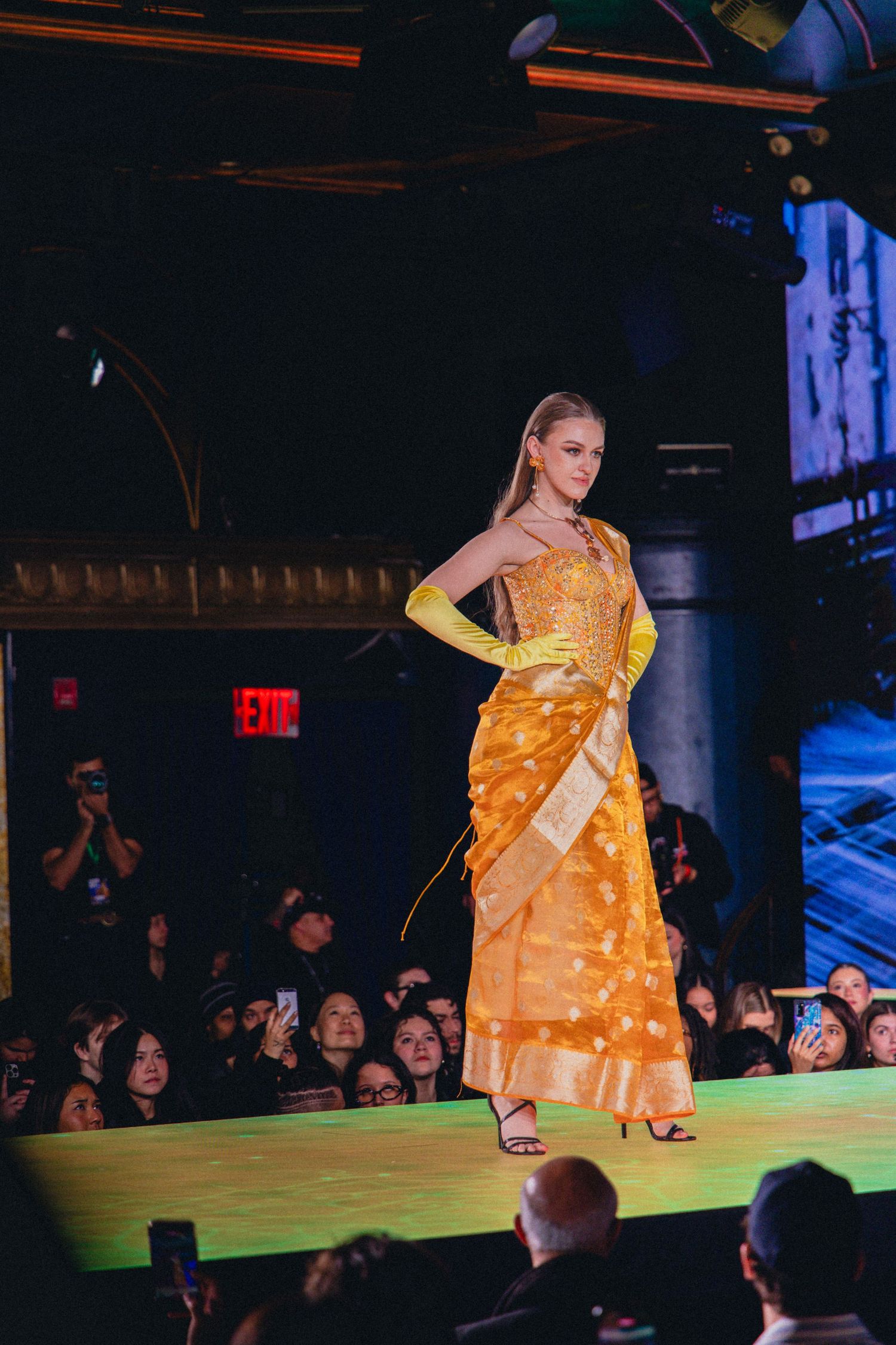 Model walks runway in flowing yellow dress with white polka dots during fashion show event.