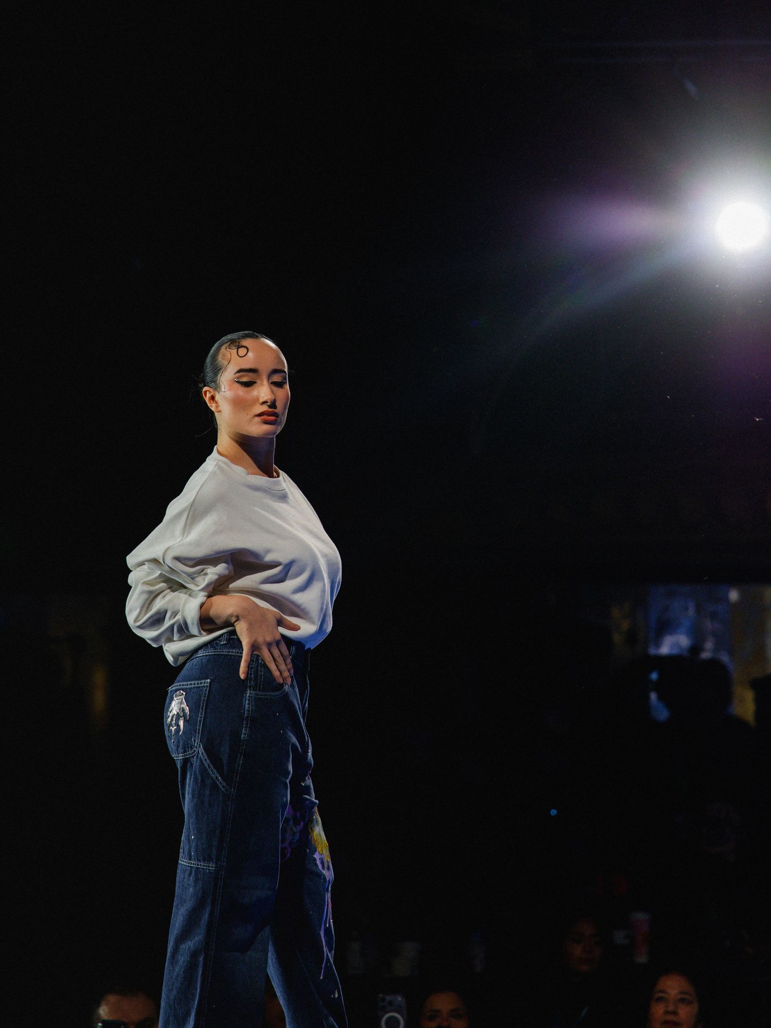 Dancer in blue jeans and light sweater performs expressive contemporary dance moves against dark background.