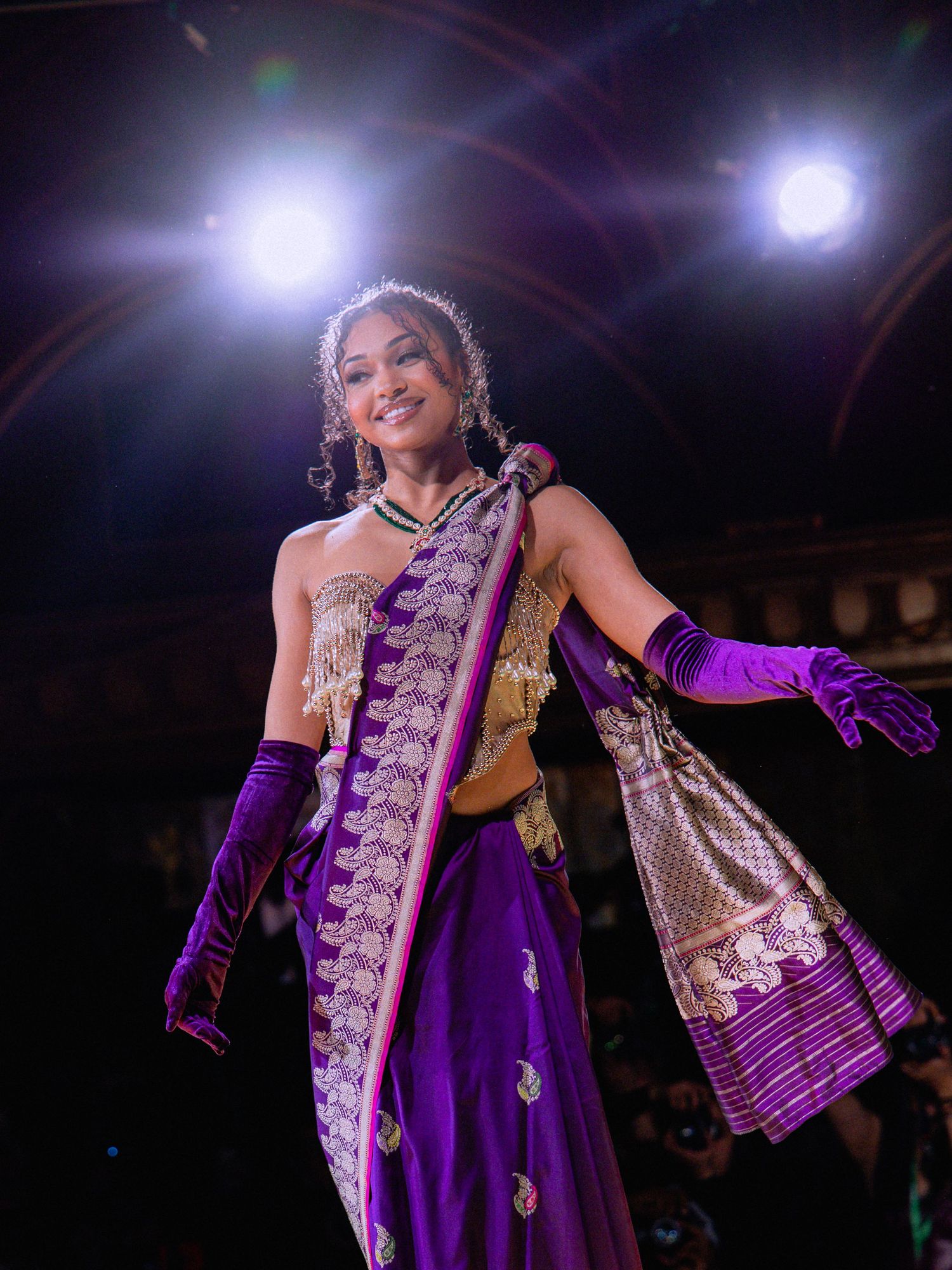 A performer in a purple gown and silver accents strikes dramatic poses under stage lighting during a theatrical show.