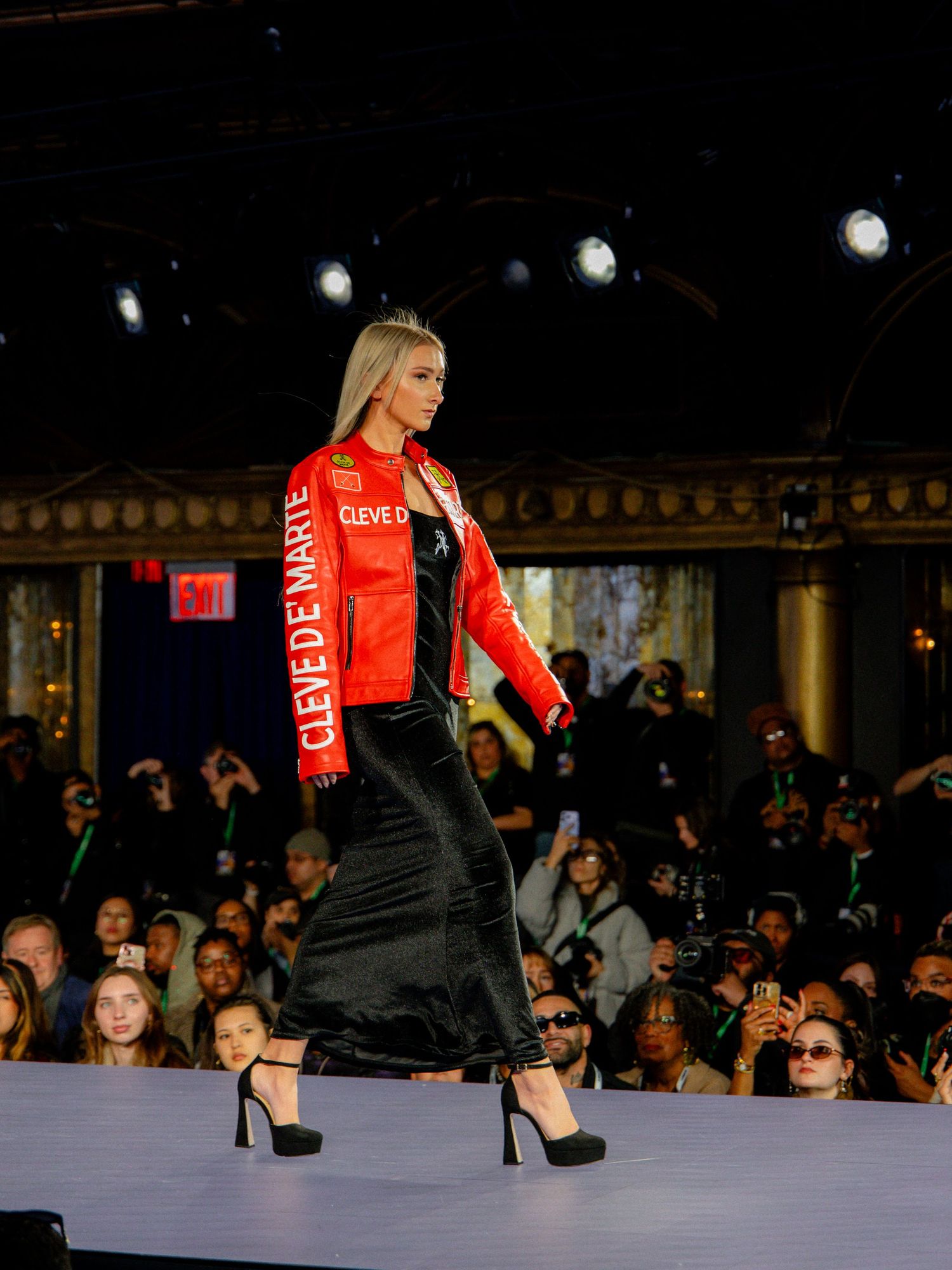 A fashion show performer in a red and black outfit walks down a runway while audience members watch in the background.