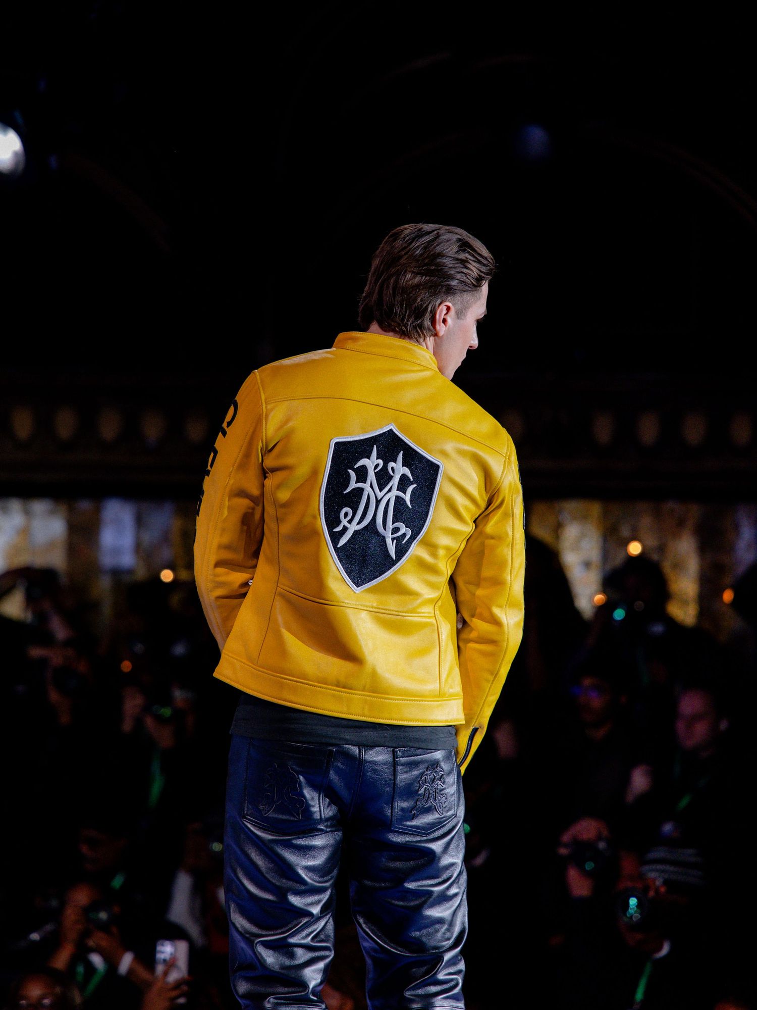 Yellow leather jacket with dark jeans shown from behind, displaying a shield logo design on the back.