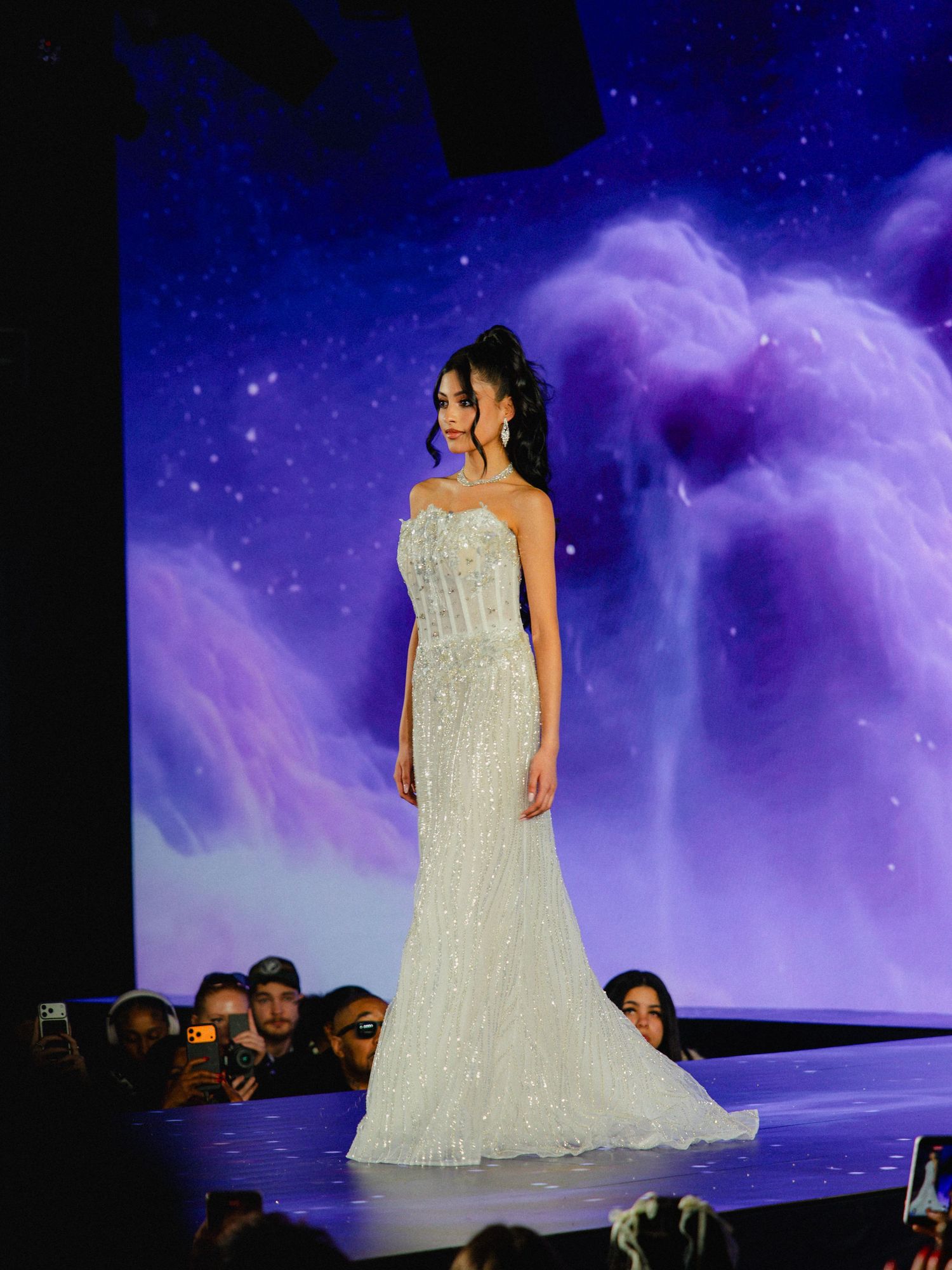 A model walks the runway in a stunning white beaded strapless wedding gown against a dreamy purple starlit backdrop.