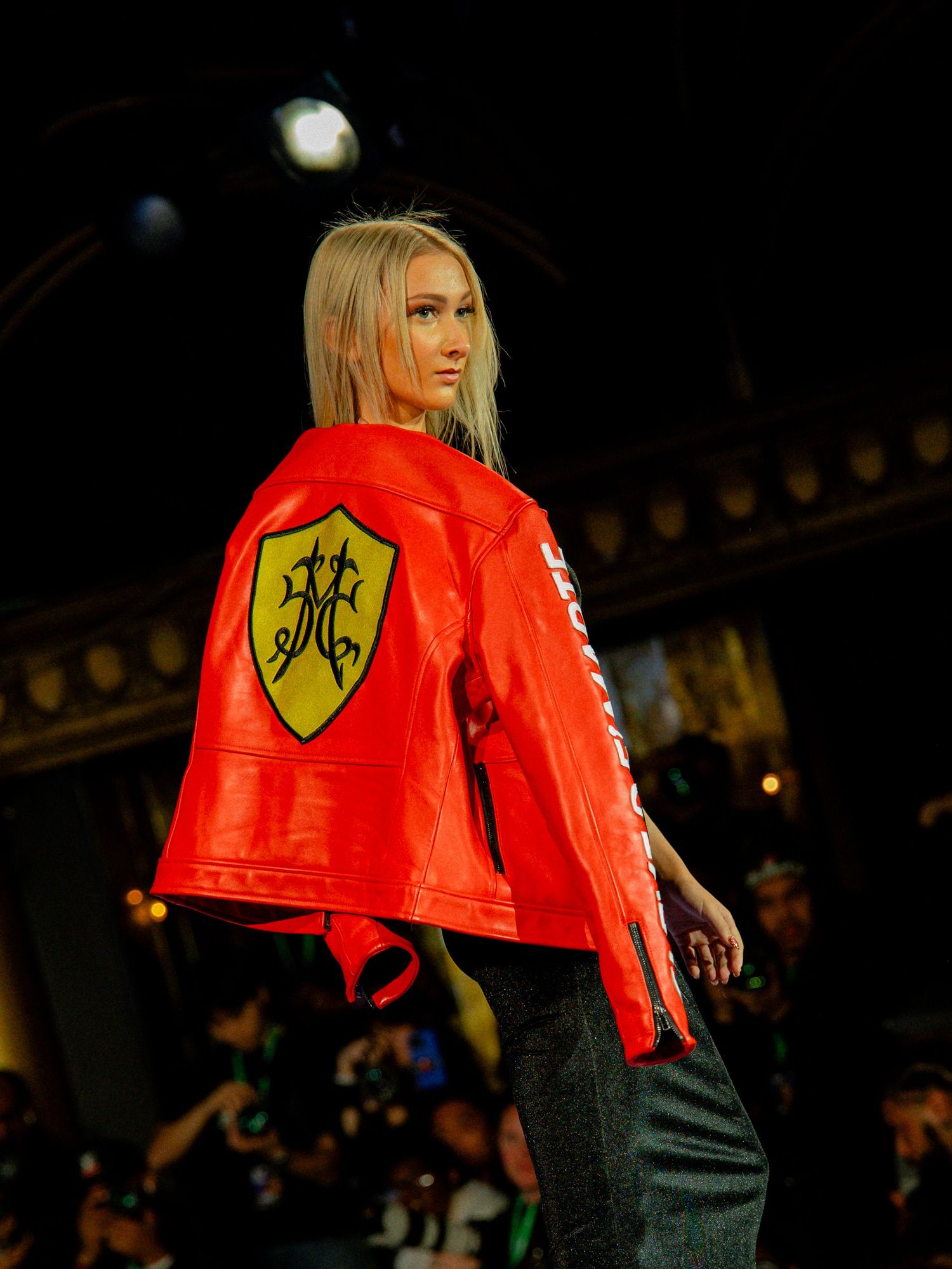 Model walks runway in bright red puffy jacket with yellow shield logo and dark pants during fashion show.