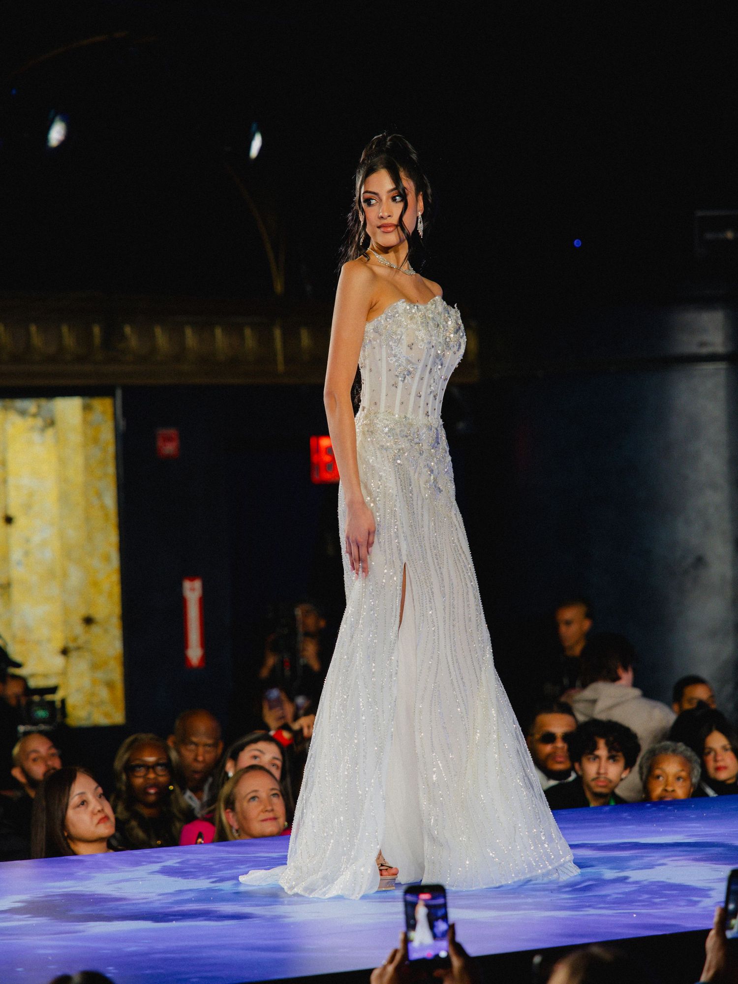 A model walks the runway in a flowing white beaded wedding gown with strapless bodice on an illuminated blue stage.