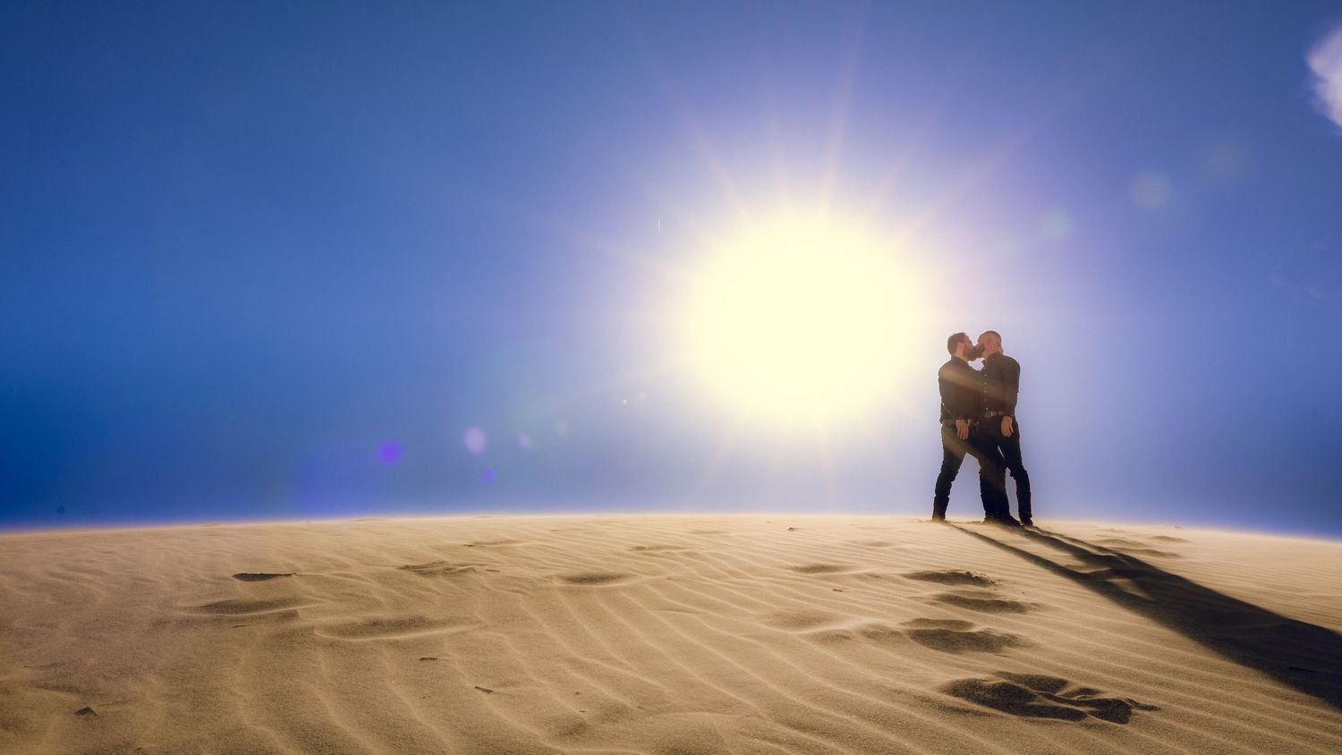 Silhouetted couple embracing on sand dune at sunset with bright sun in background.