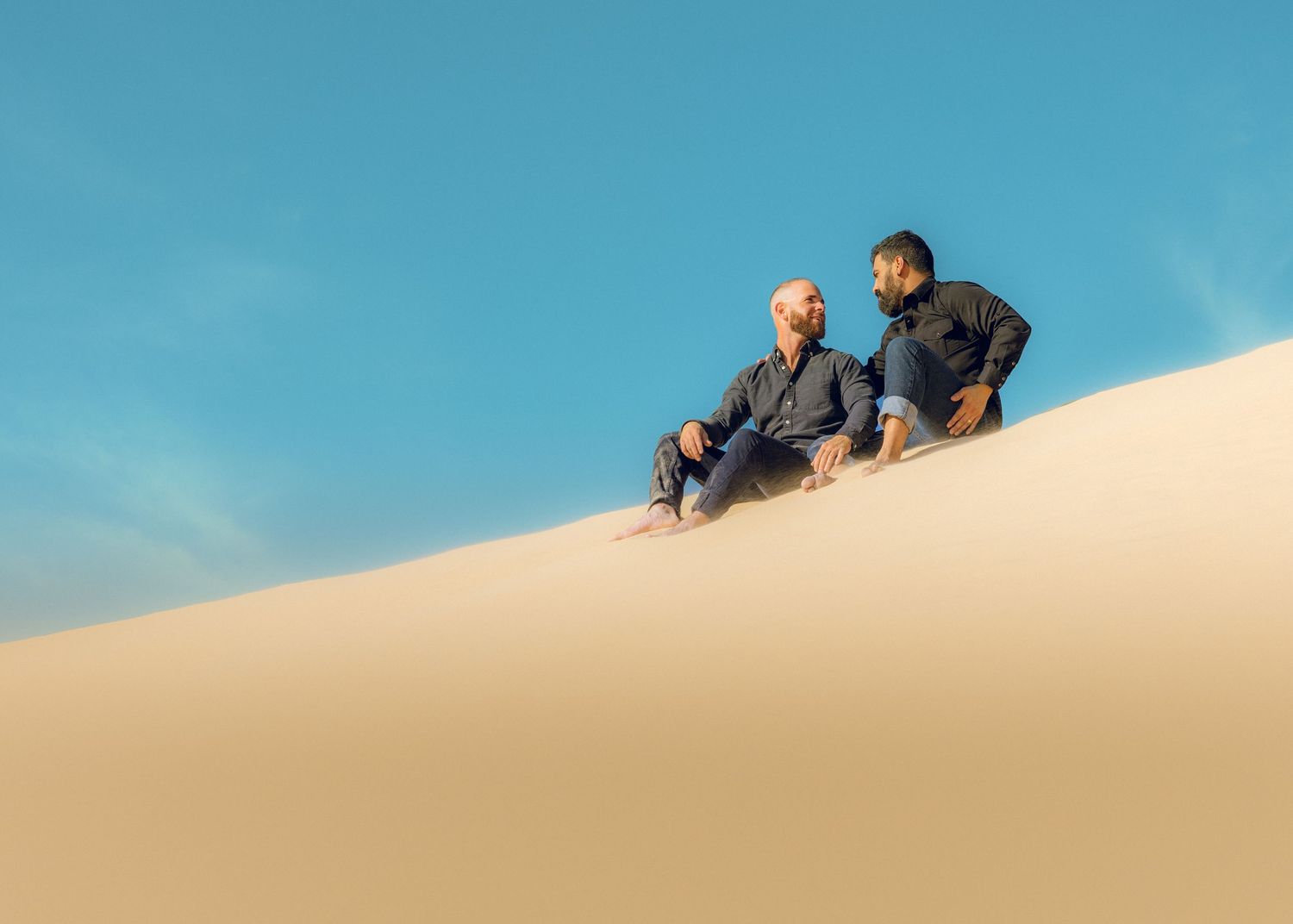 People climbing and playing on large sand dunes under blue sky.