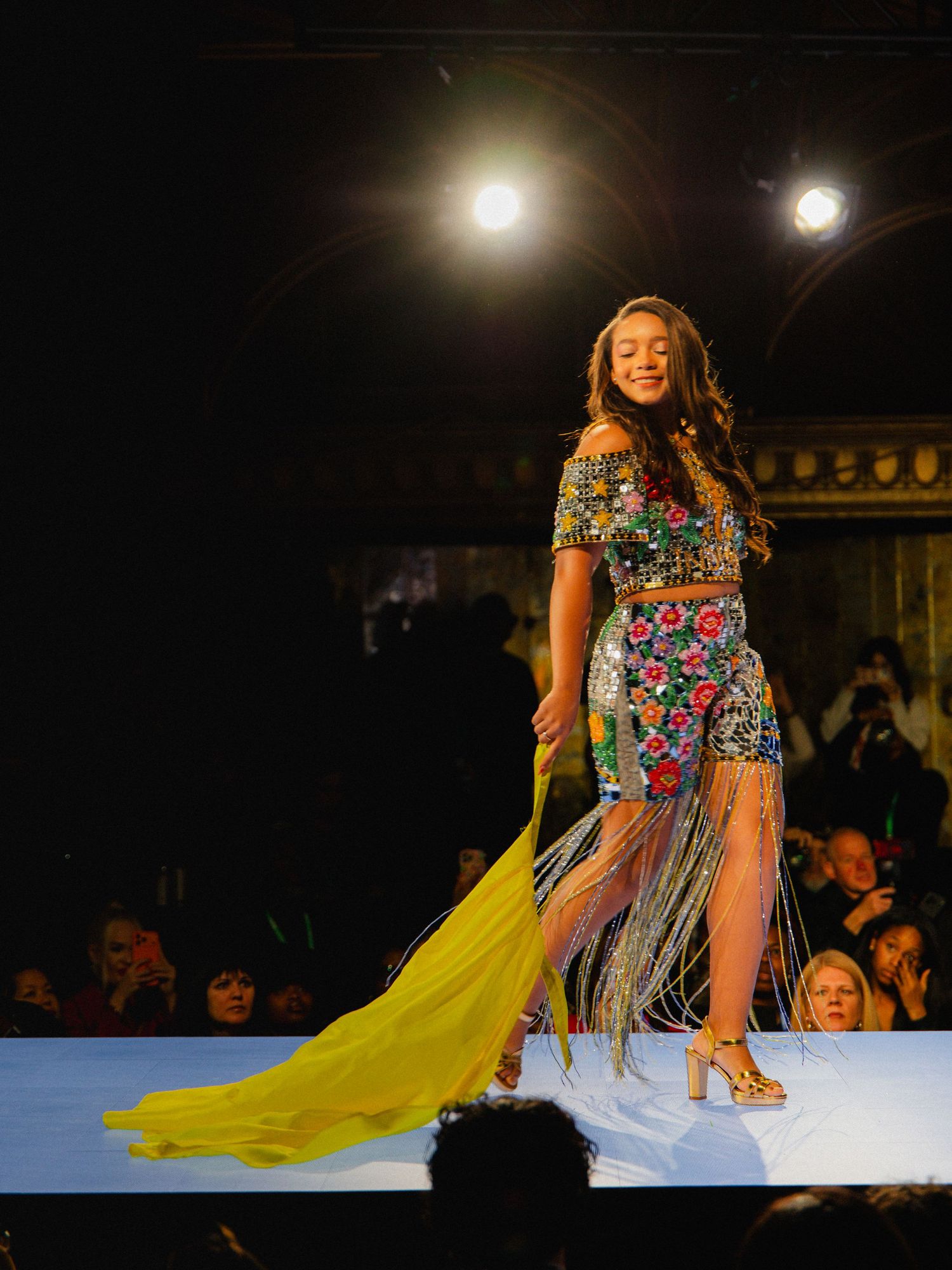 Model walks runway in colorful floral print dress with flowing yellow train at fashion show under dramatic lighting.
