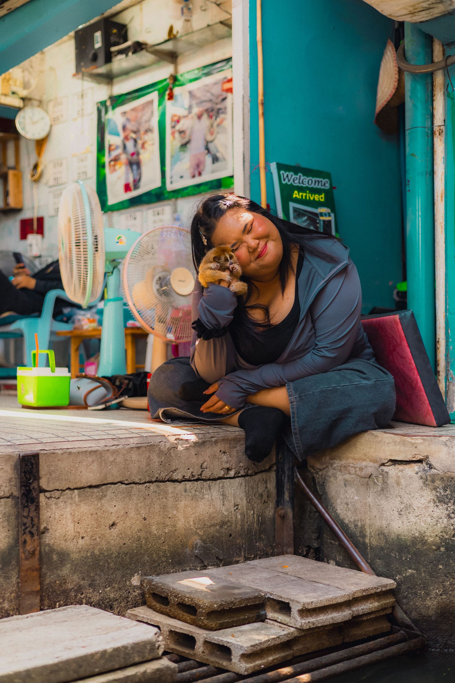 Woman sitting on a step outside a shop, holding a small dog, with a fan and teal wall in the background.