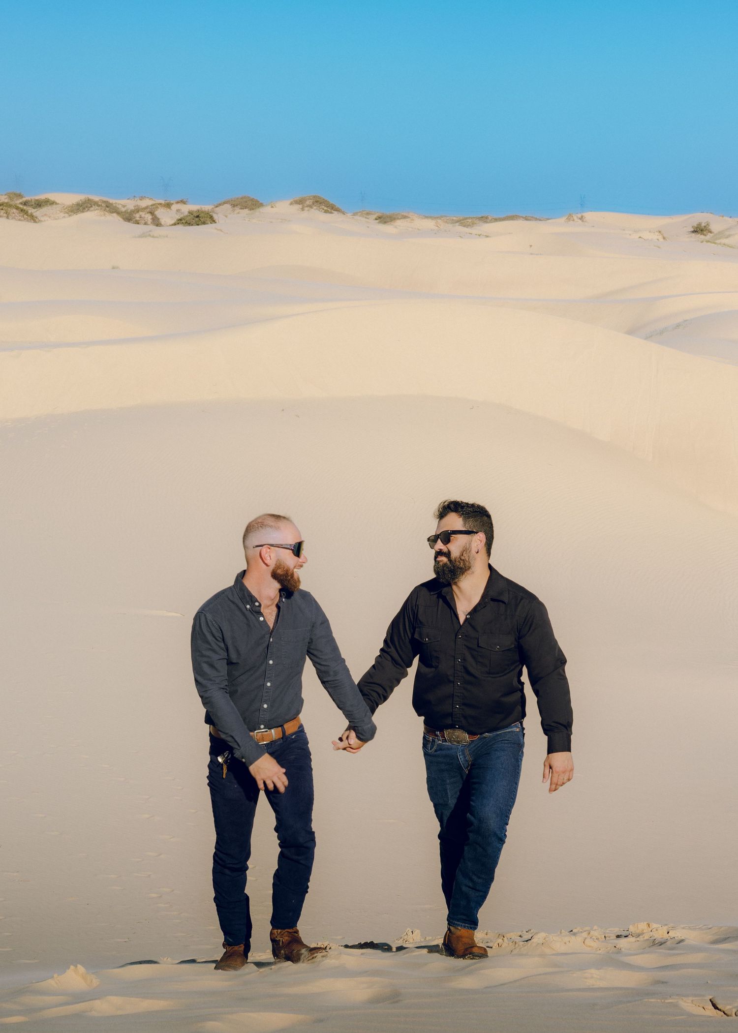Two people walking hand in hand across sandy dunes under bright blue sky.
