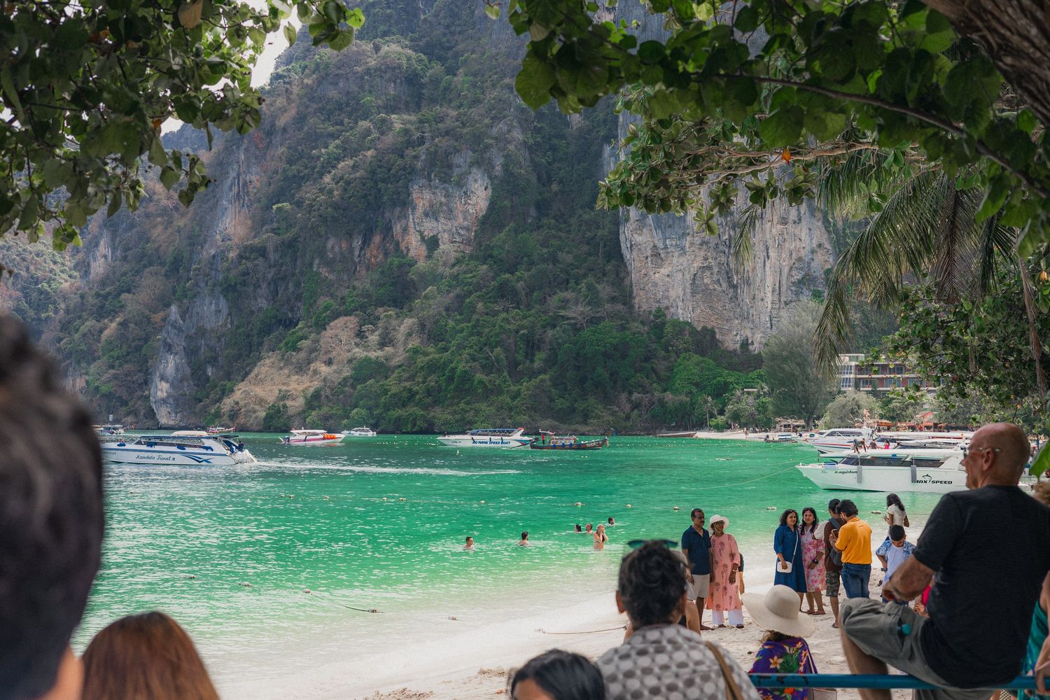 Tourists on a white sandy beach with turquoise water, boats, and dramatic limestone cliffs in Koh Phi Phi, Thailand.