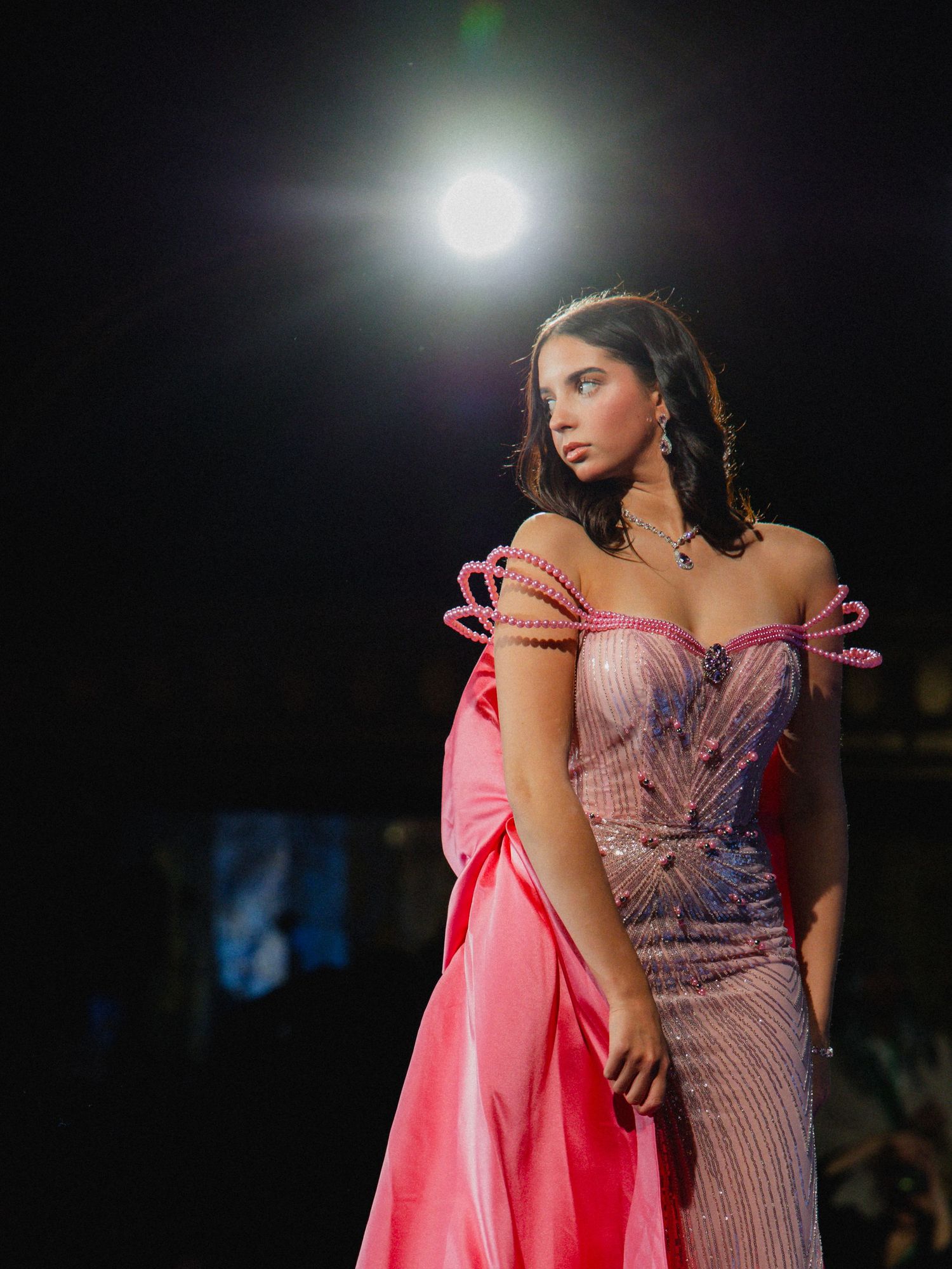 Model wearing a glamorous pink and silver evening gown with off-shoulder design under dramatic stage lighting.