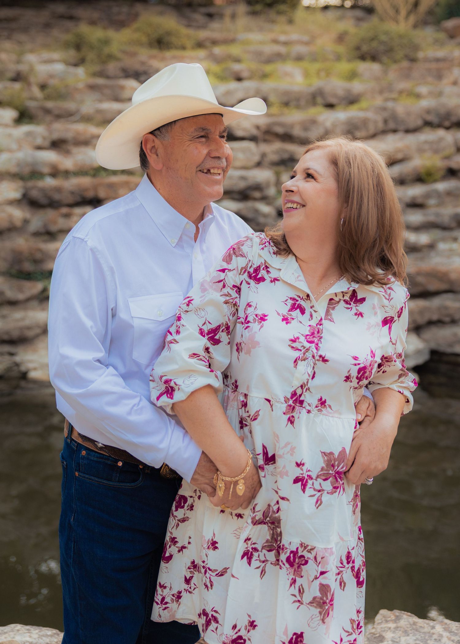 Romantic couple embracing by a lake, with woman wearing floral dress and man in western attire.