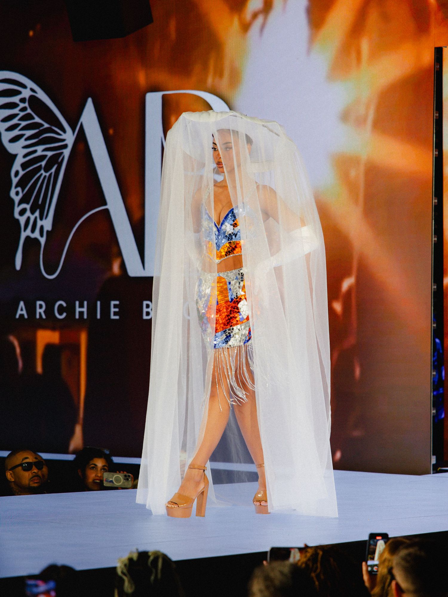 A fashion model walks the runway in a flowing white veil and colorful floral dress during a glamorous evening show.