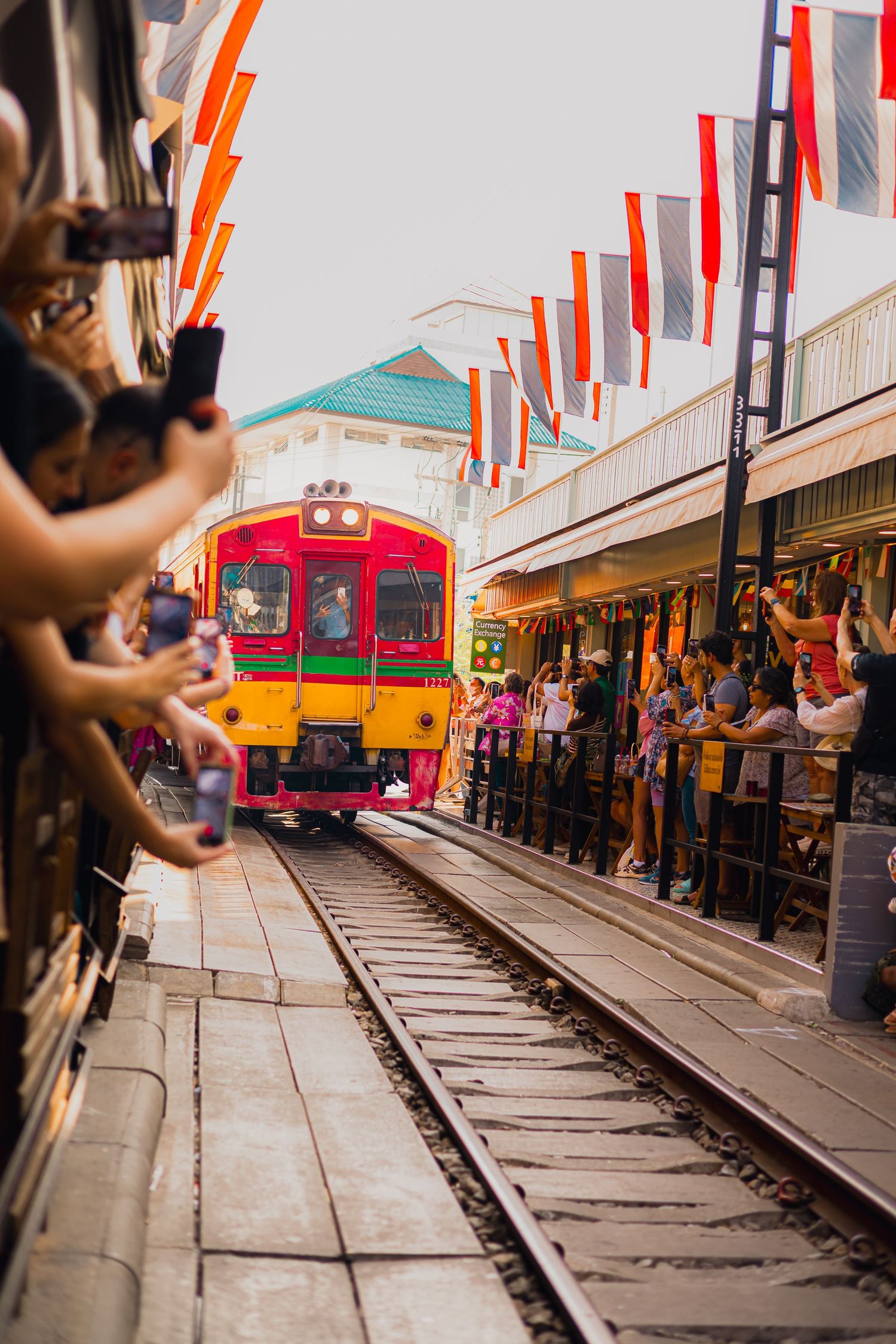 Colorful red and yellow train arrives at busy Mae Klong Railway Market as tourists photograph it.