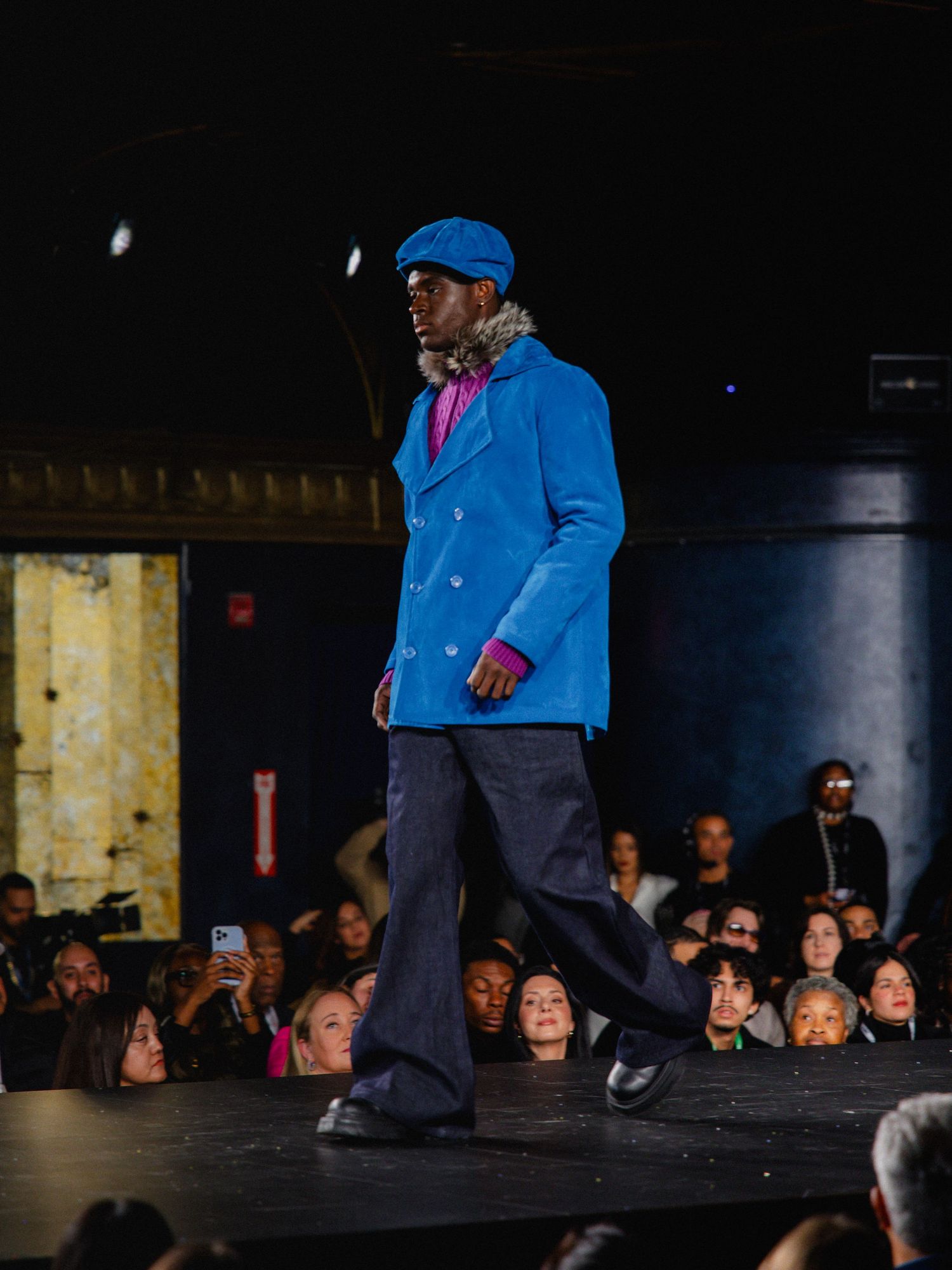 A performer in a bright blue jacket and dark pants walks across a dimly lit stage with an audience visible in the background.