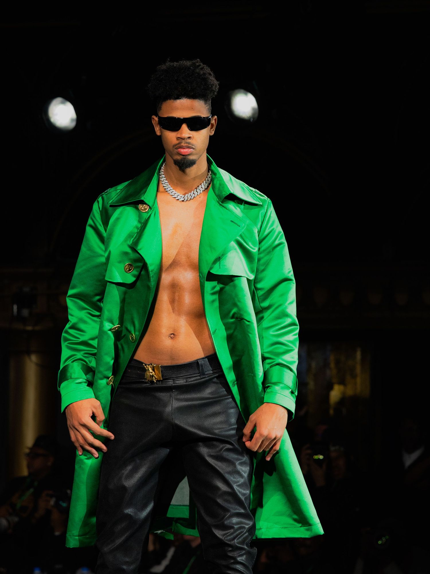 Fashion model wearing bright green blazer with black pants and sunglasses poses on dark runway.