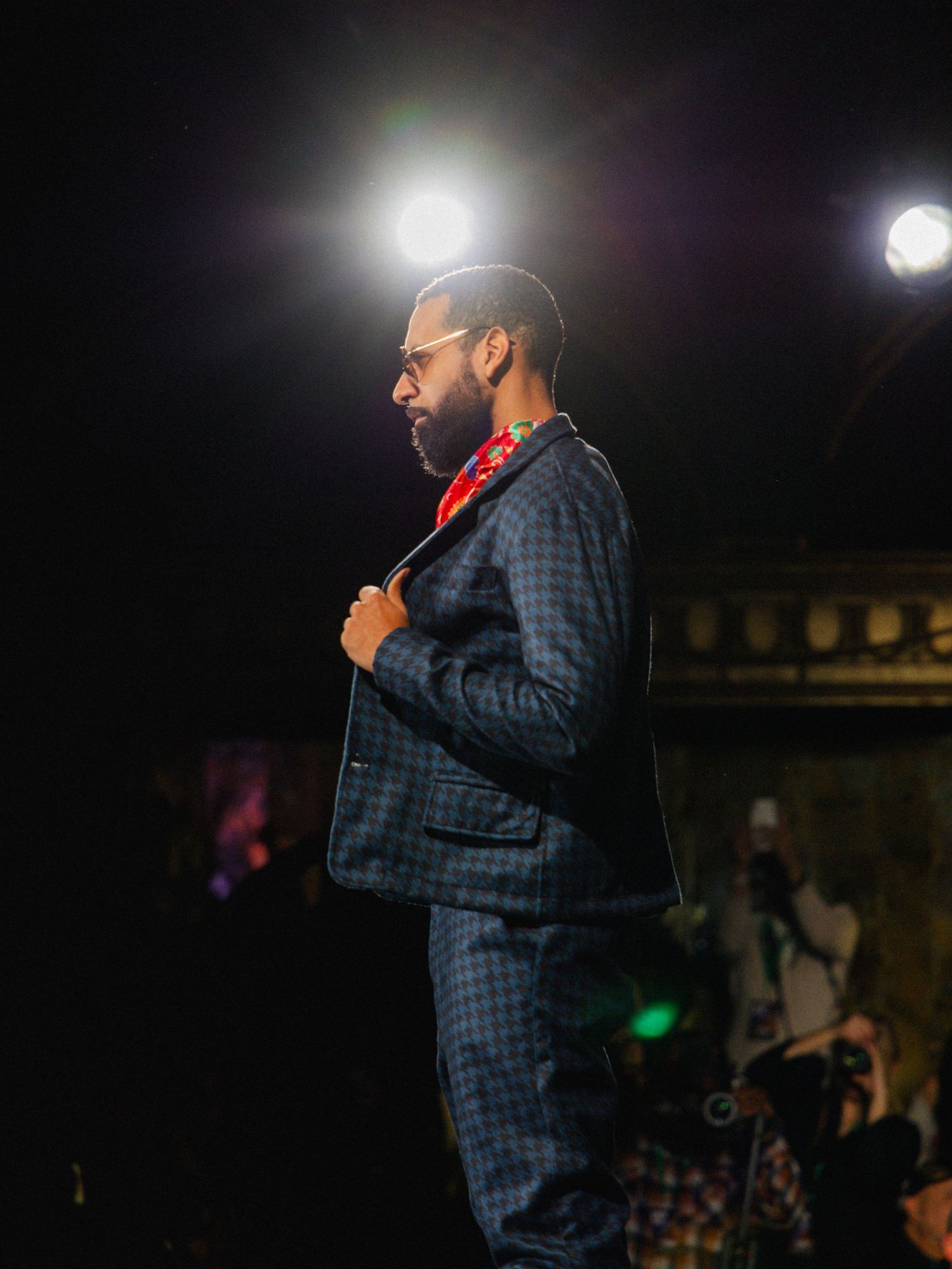 A performer in a plaid suit stands on stage under bright spotlights during a nighttime show.