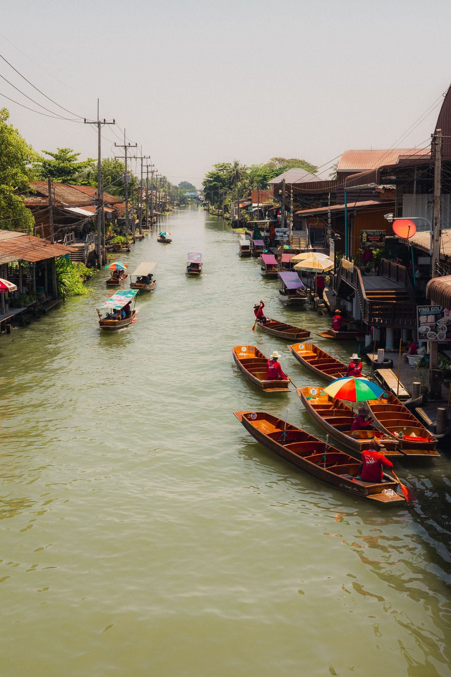 Wooden boats navigate a bustling floating market canal in Thailand, lined with traditional stilt houses and vendors.