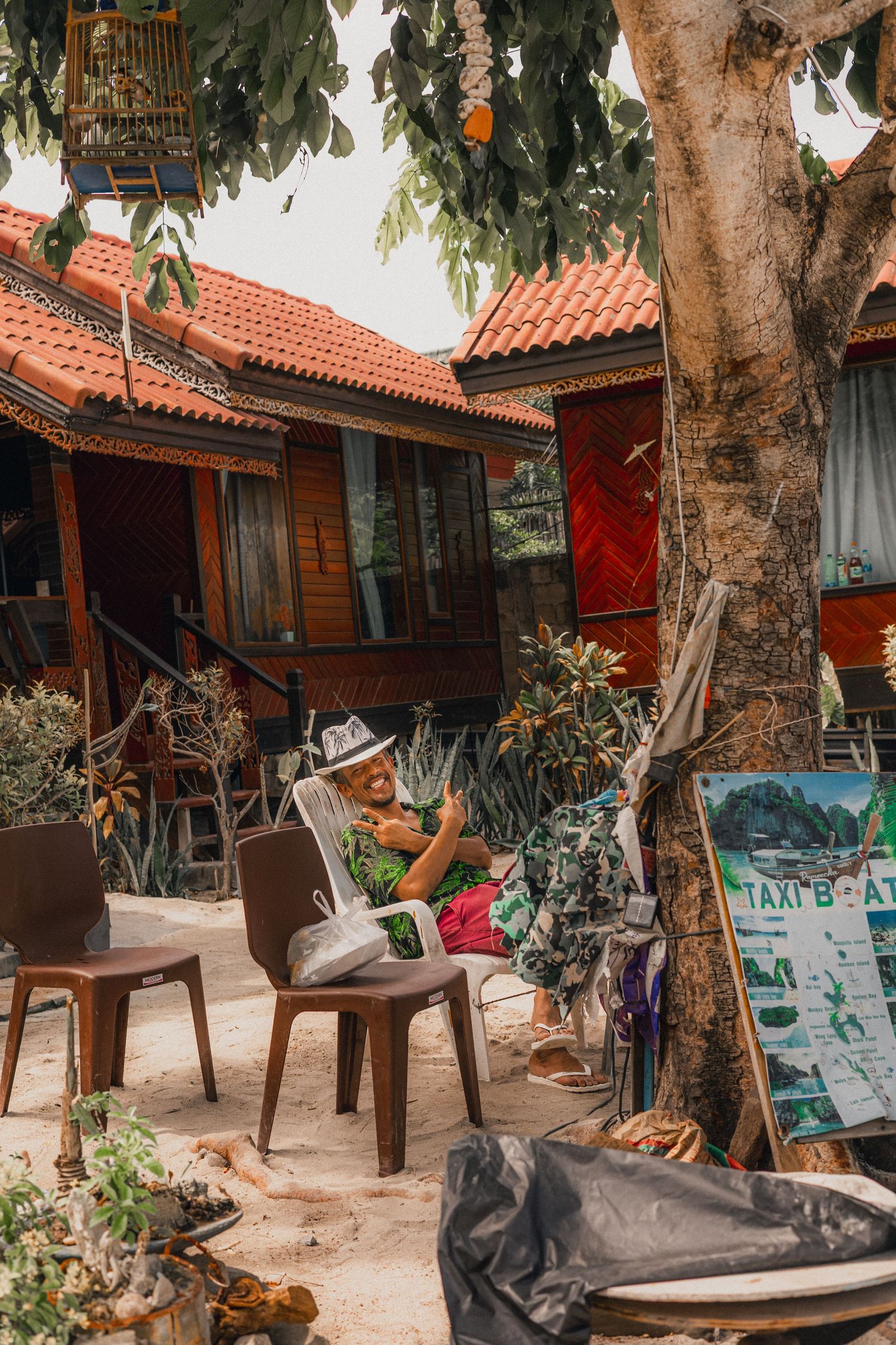 Man relaxing in chair reading newspaper outside rustic wooden bungalow with red tiled roof in tropical setting.