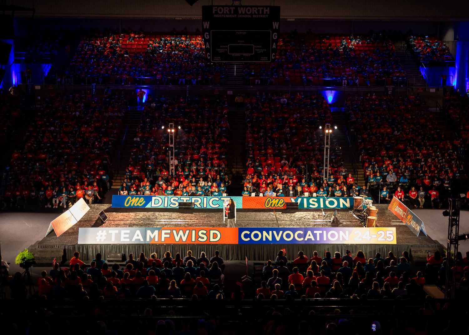 Large indoor auditorium filled with audience members under blue and red lighting.