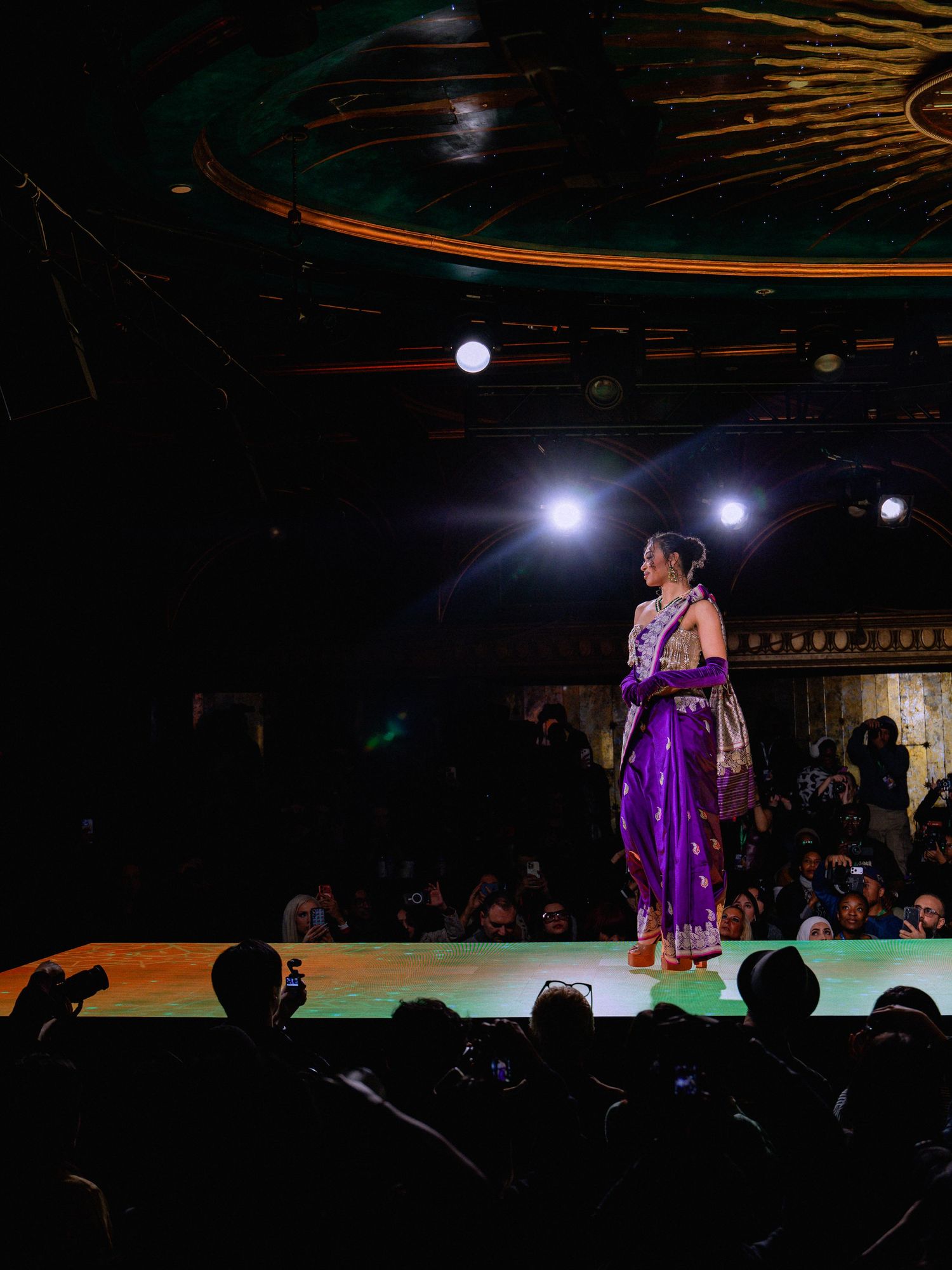 Fashion models walk the runway in purple sequined dresses under dramatic stage lighting during an evening show.