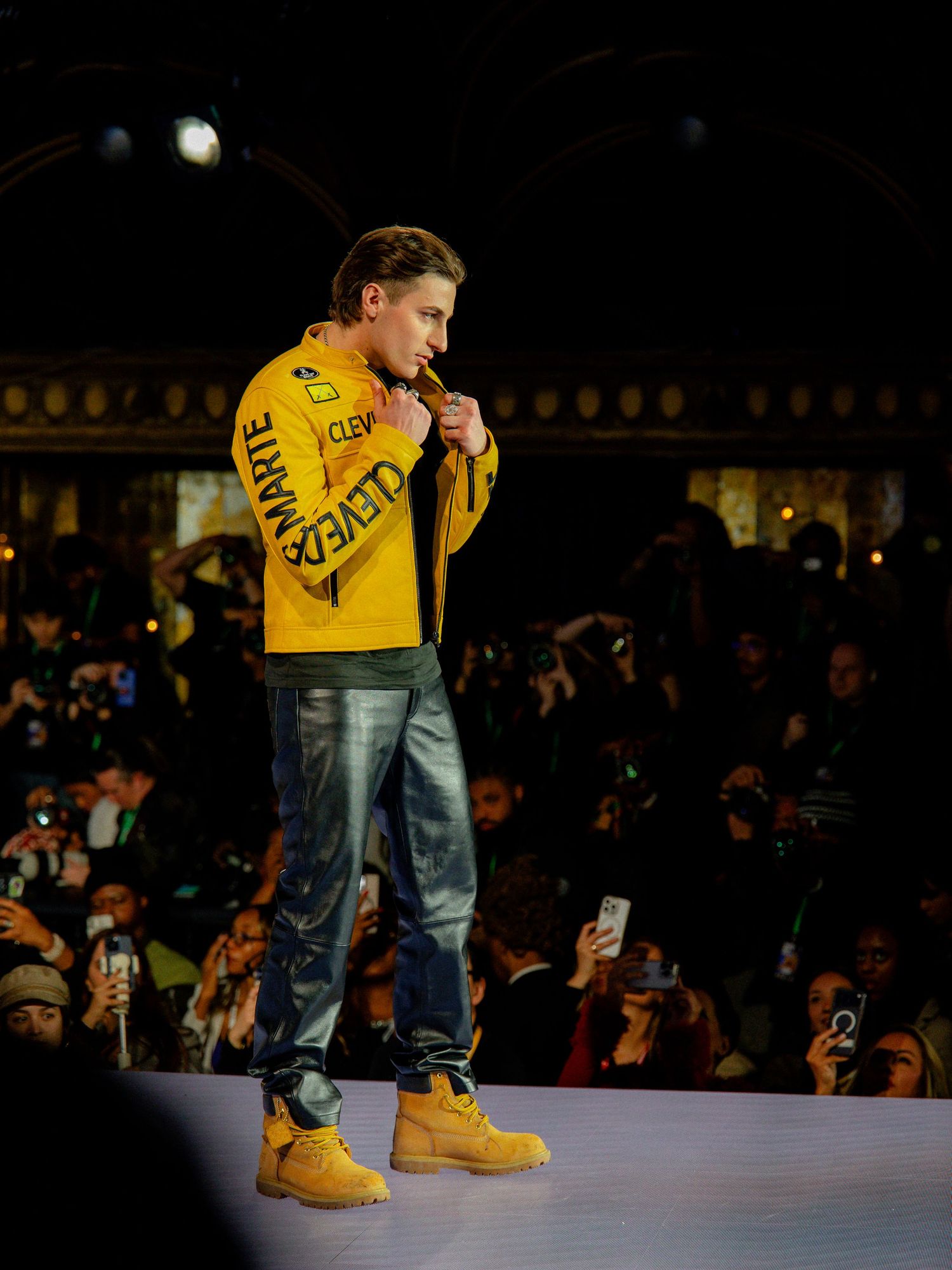 A performer in a yellow jacket and jeans stands on a stage during a live performance with a dark audience background.