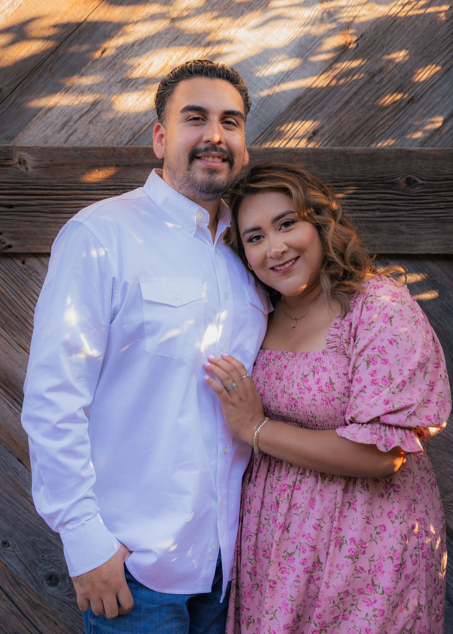 Couple in pink dress and white shirt pose together on wooden deck during sunset.