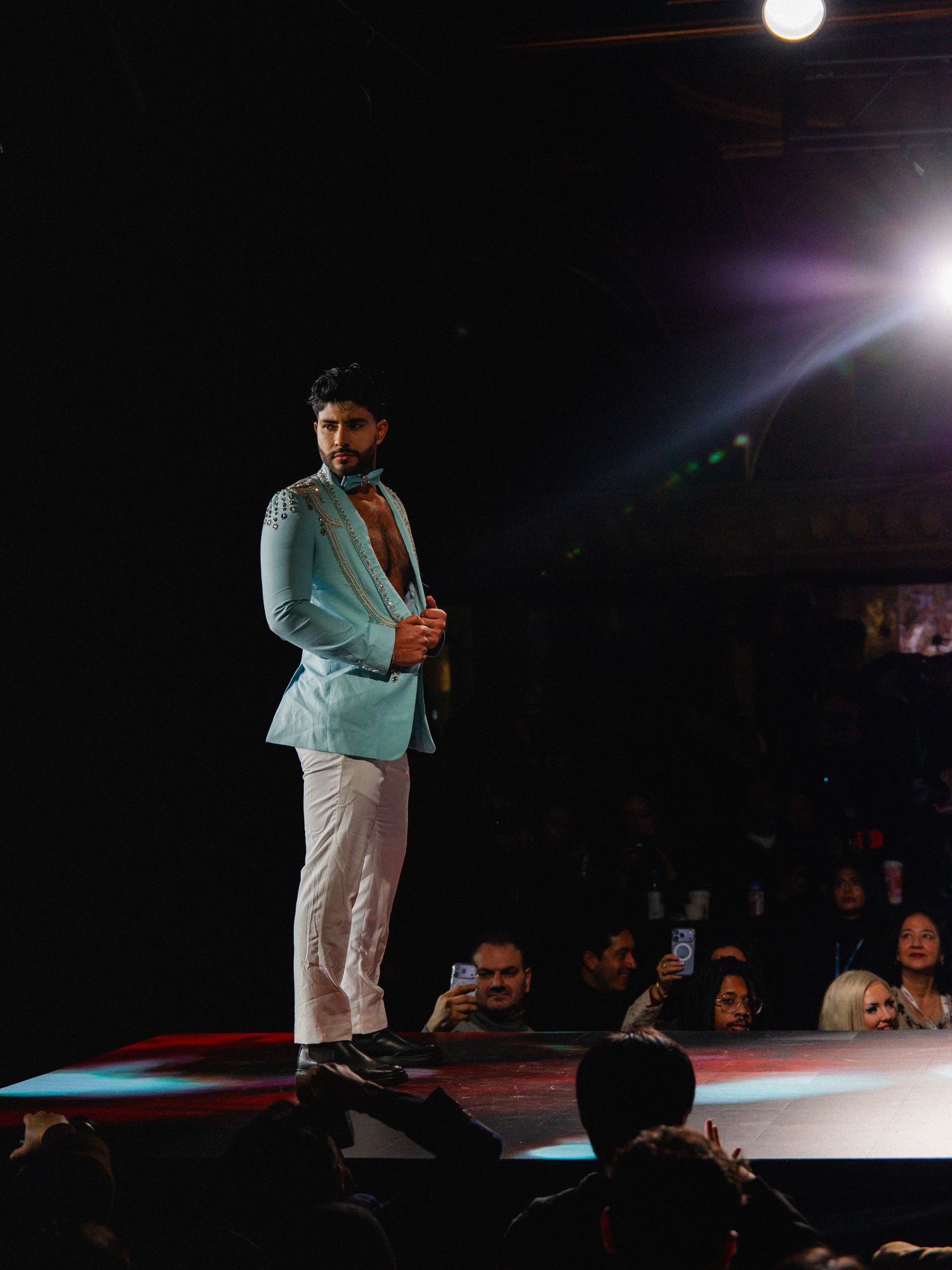 A performer in light blue jacket and white pants stands on stage under dramatic spotlighting during a show.