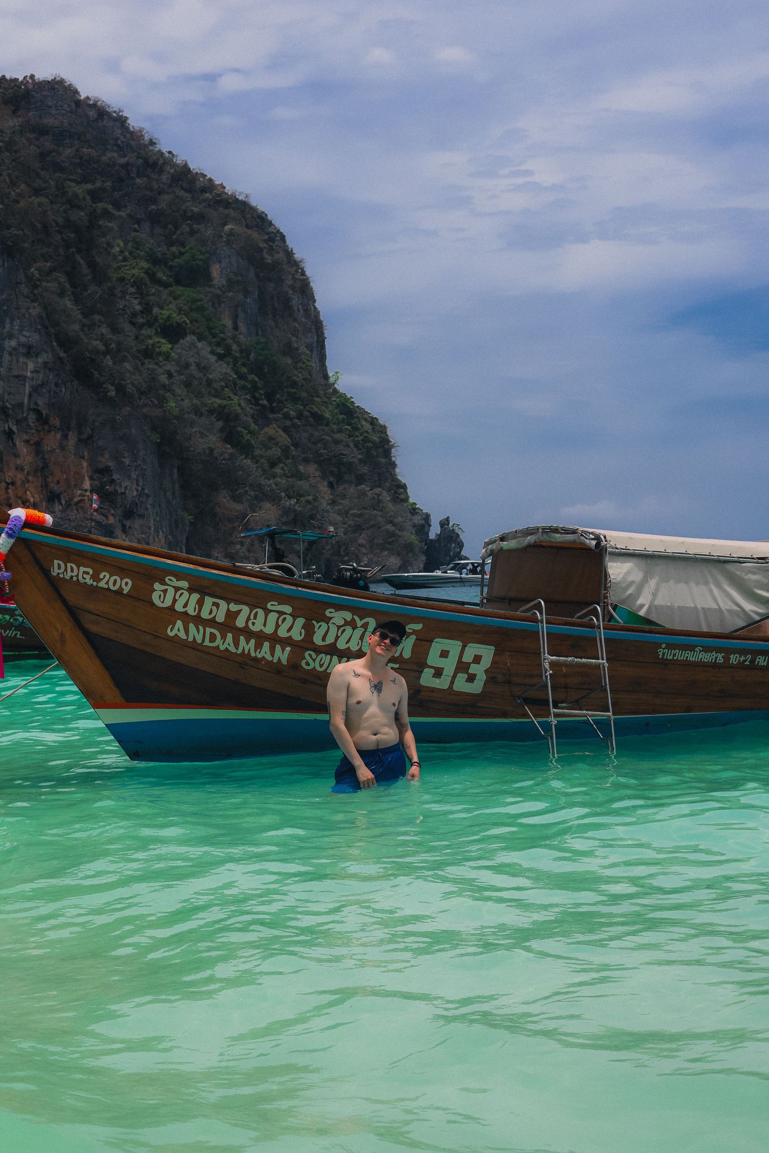 Man standing in turquoise waters beside a traditional Thai longtail boat near limestone cliffs in Koh Phi Phi, Thailand.
