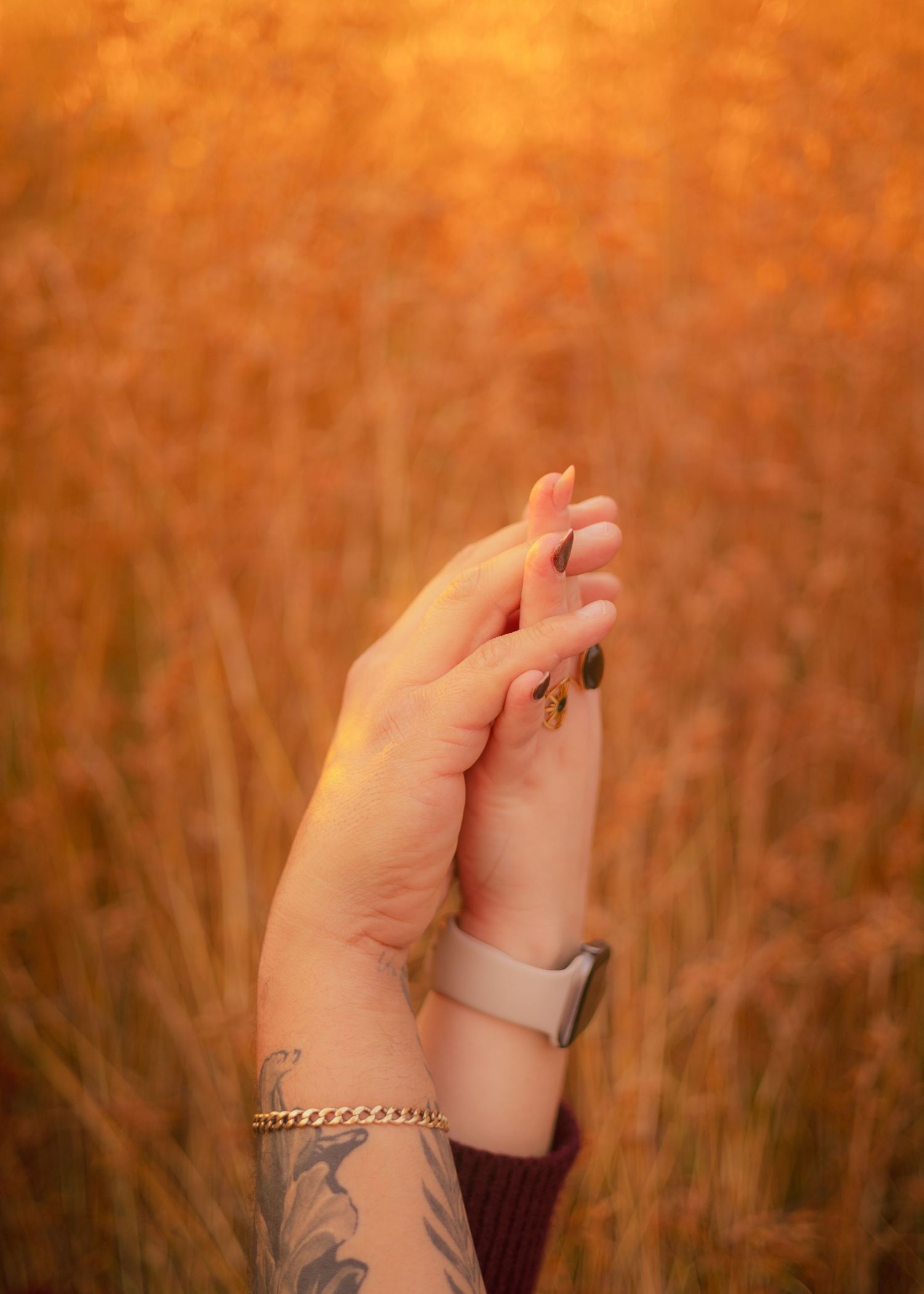 Hands with tattoo and bracelet reaching upward against warm orange sunset in wheat field.