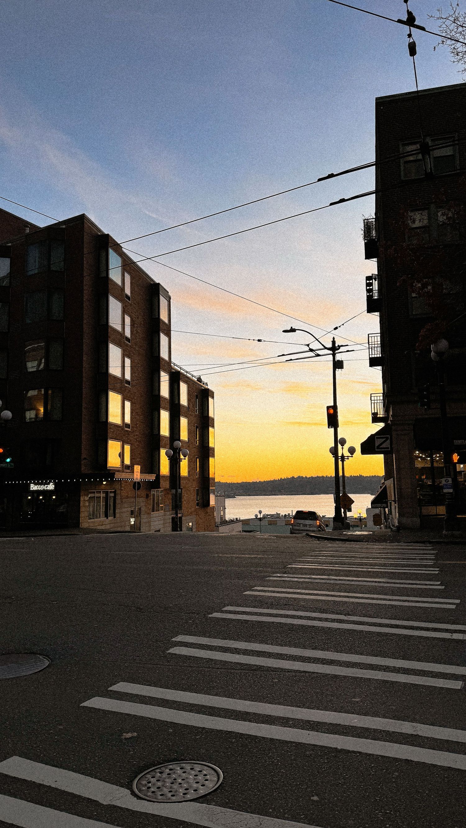 Empty city street with crosswalk and buildings silhouetted against golden sunset sky.