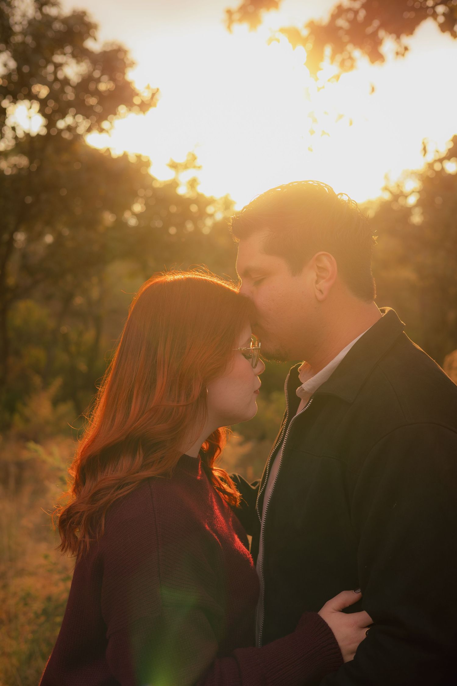 A couple shares an intimate moment at sunset with golden light filtering through trees in the background.